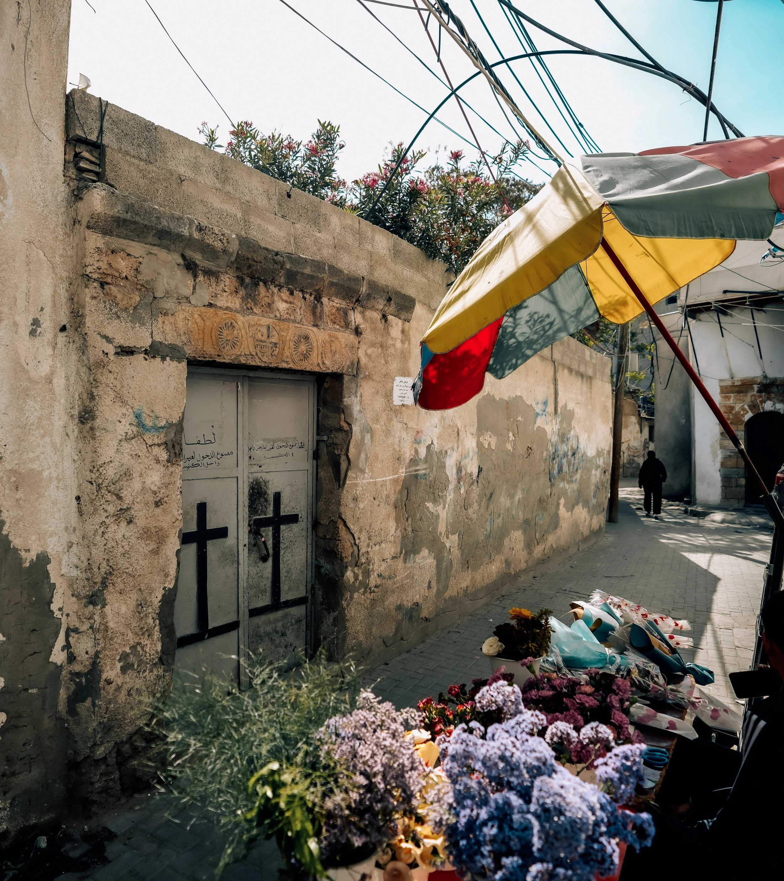 Flowers laying on the street with a wall and a building in the background, and a person walking away in the distance.