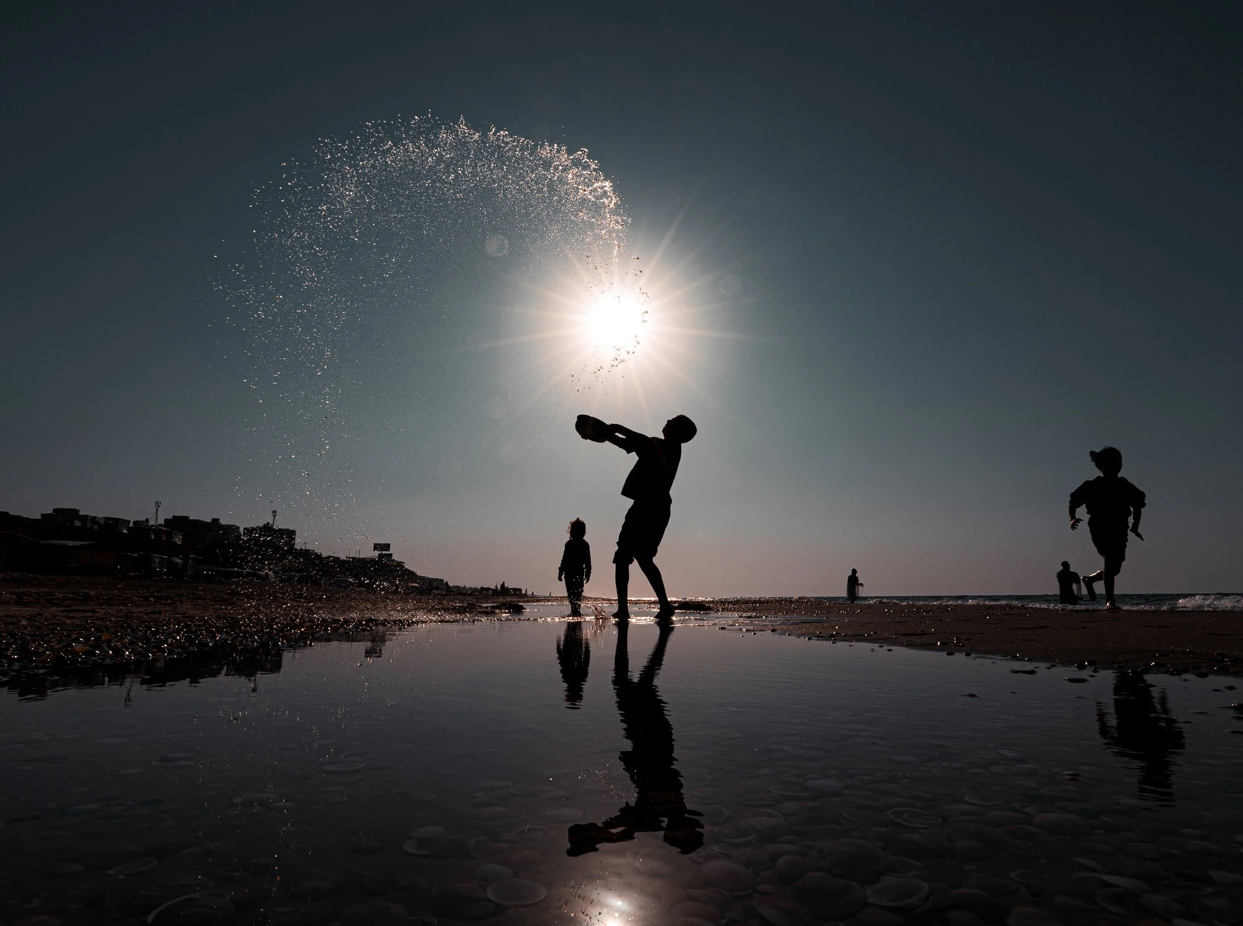 Silhouettes of children playing by the beach in Gaza with water splashing, during sunset with the sun shining brightly in the sky.