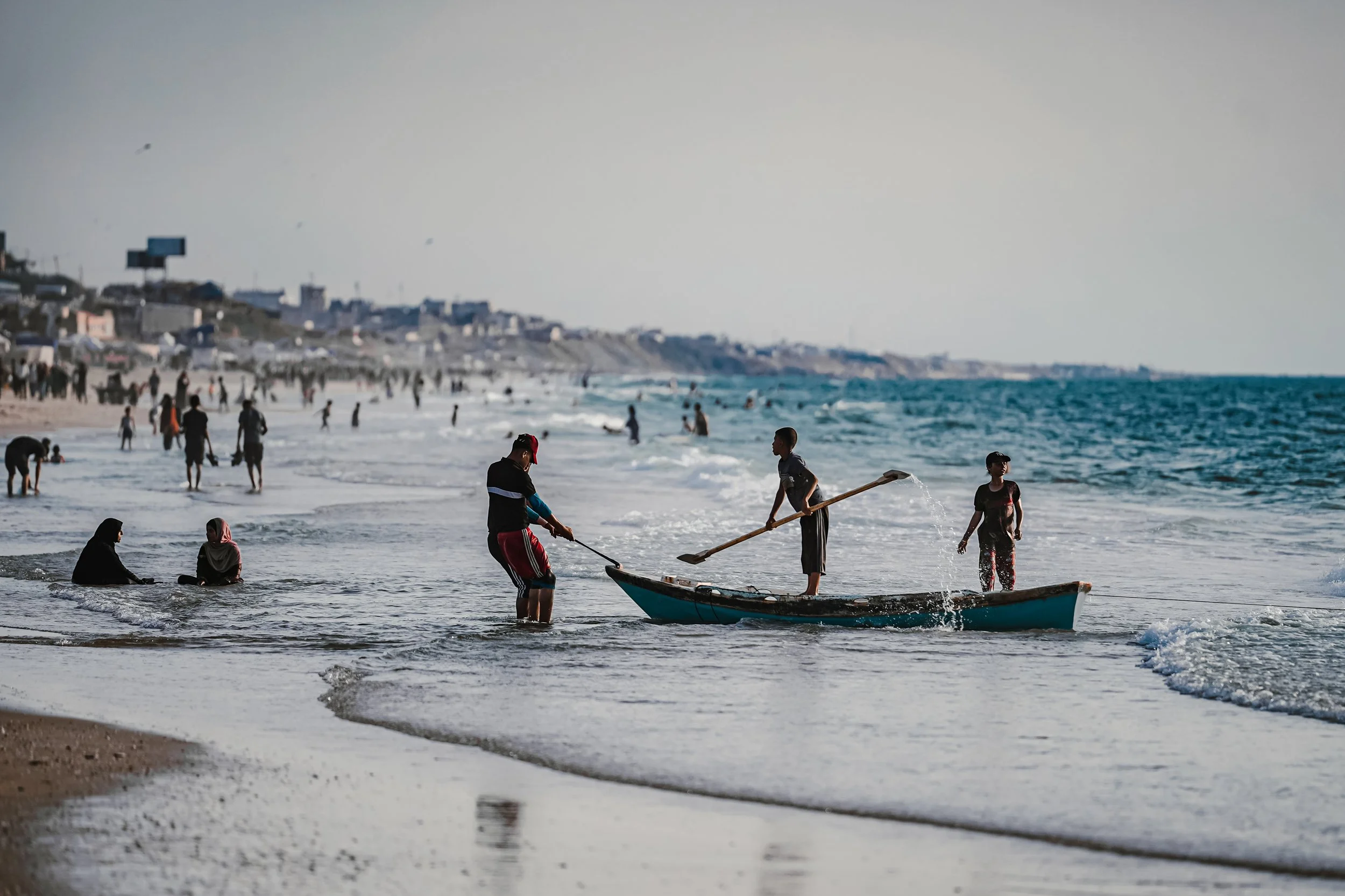 People on the beach in Gaza, some standing in water, others sitting or walking along the shore, with a boat and a small boat in the foreground, and buildings on a hillside in the background.
