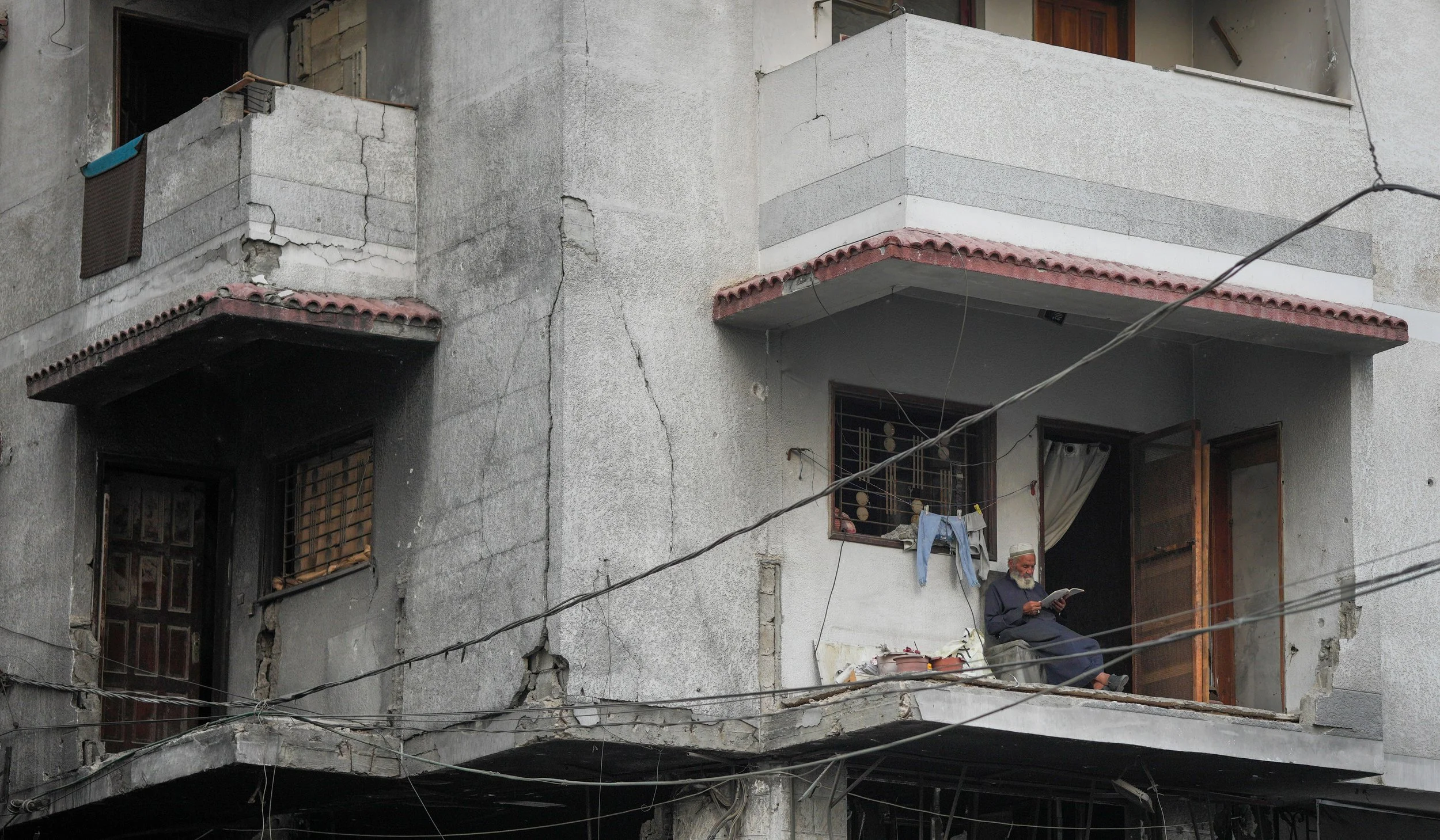 A damaged building with visible cracks and signs of wear. A man with a beard is sitting on the open, destroyed balcony reading a book.