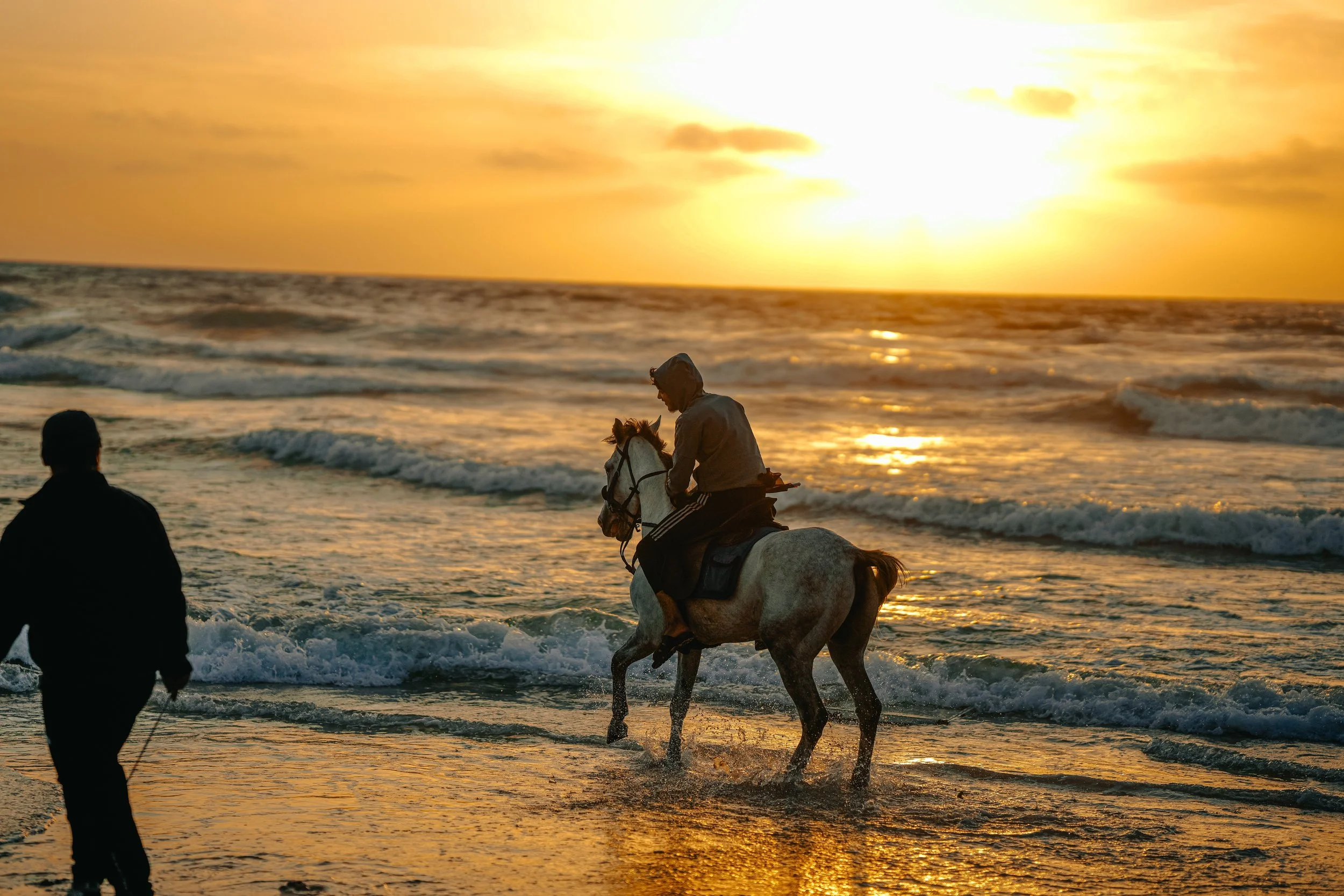 A young Palestinian man riding a horse through ocean waves during sunset, with another person standing on the beach watching.