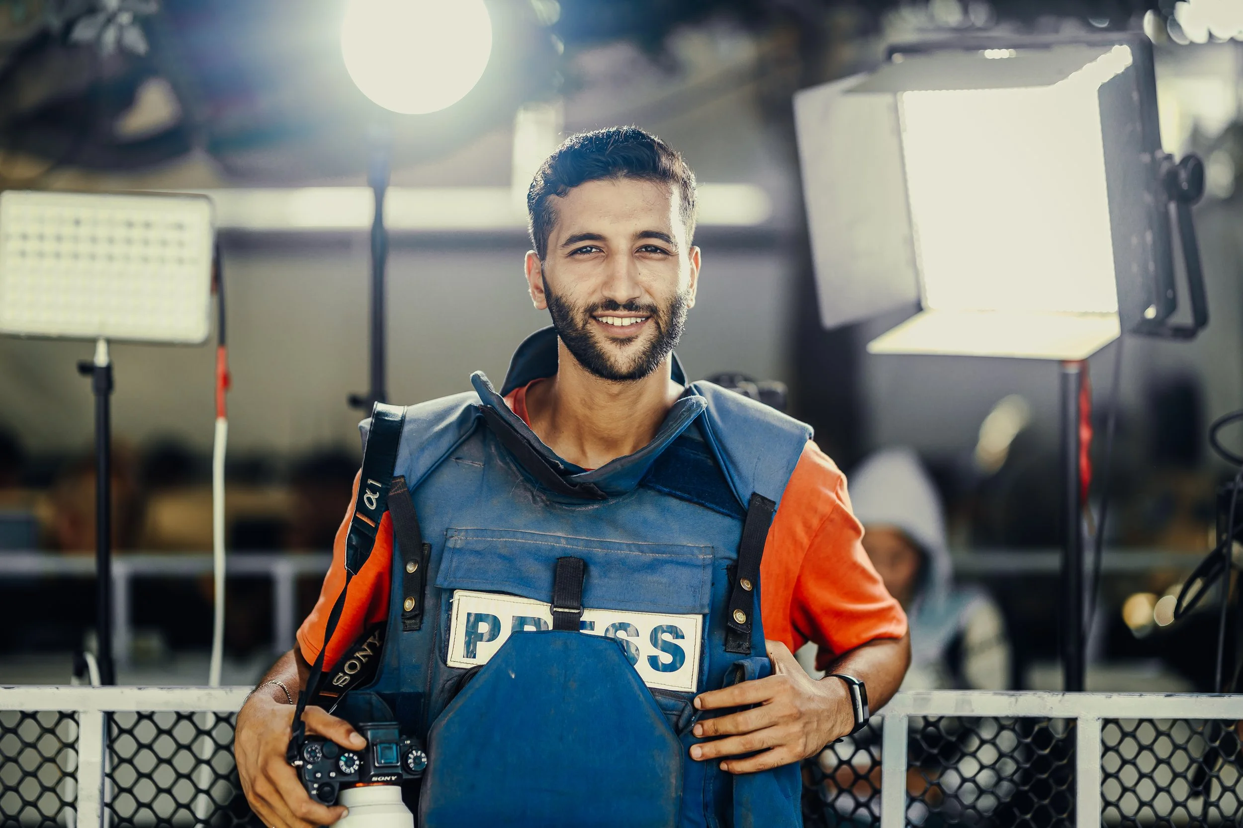 A Palestinian photojournalist, wearing a vest labeled "PRESS" and holding a camera, standing in front of light equipment.