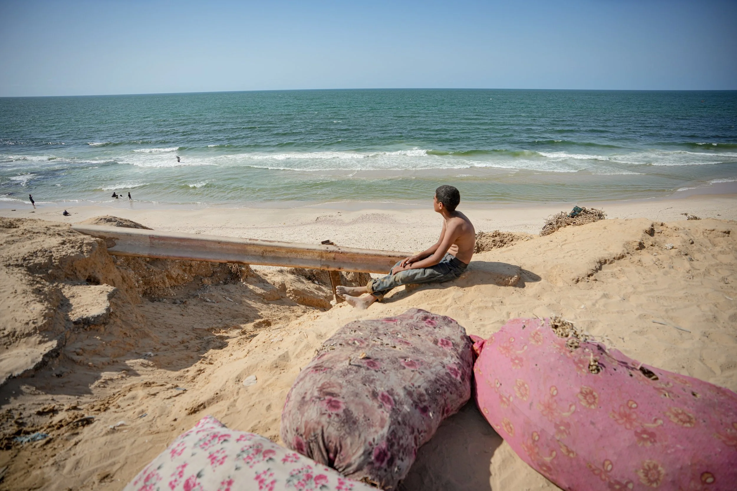 A young boy sitting on the sandy beach in Gaza with pink floral blankets, looking out at the ocean.