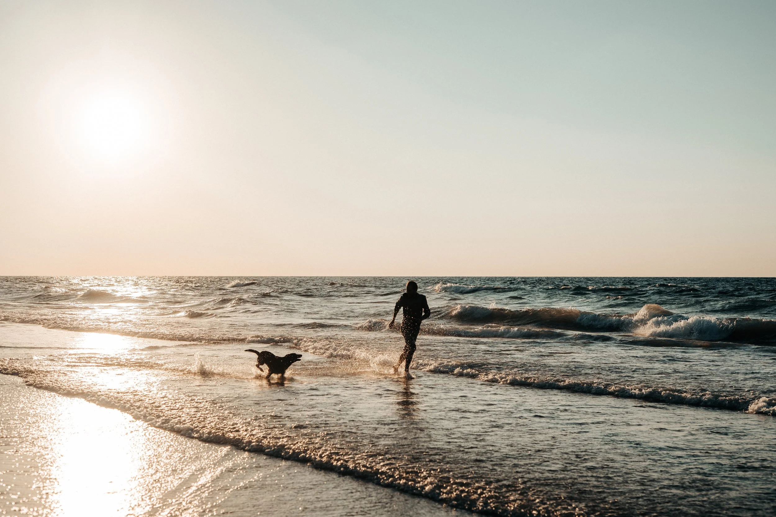 A Palestinian man and a dog running along the shoreline of the beach in Gaza during sunset, with the sun low in the sky and gentle waves around them.