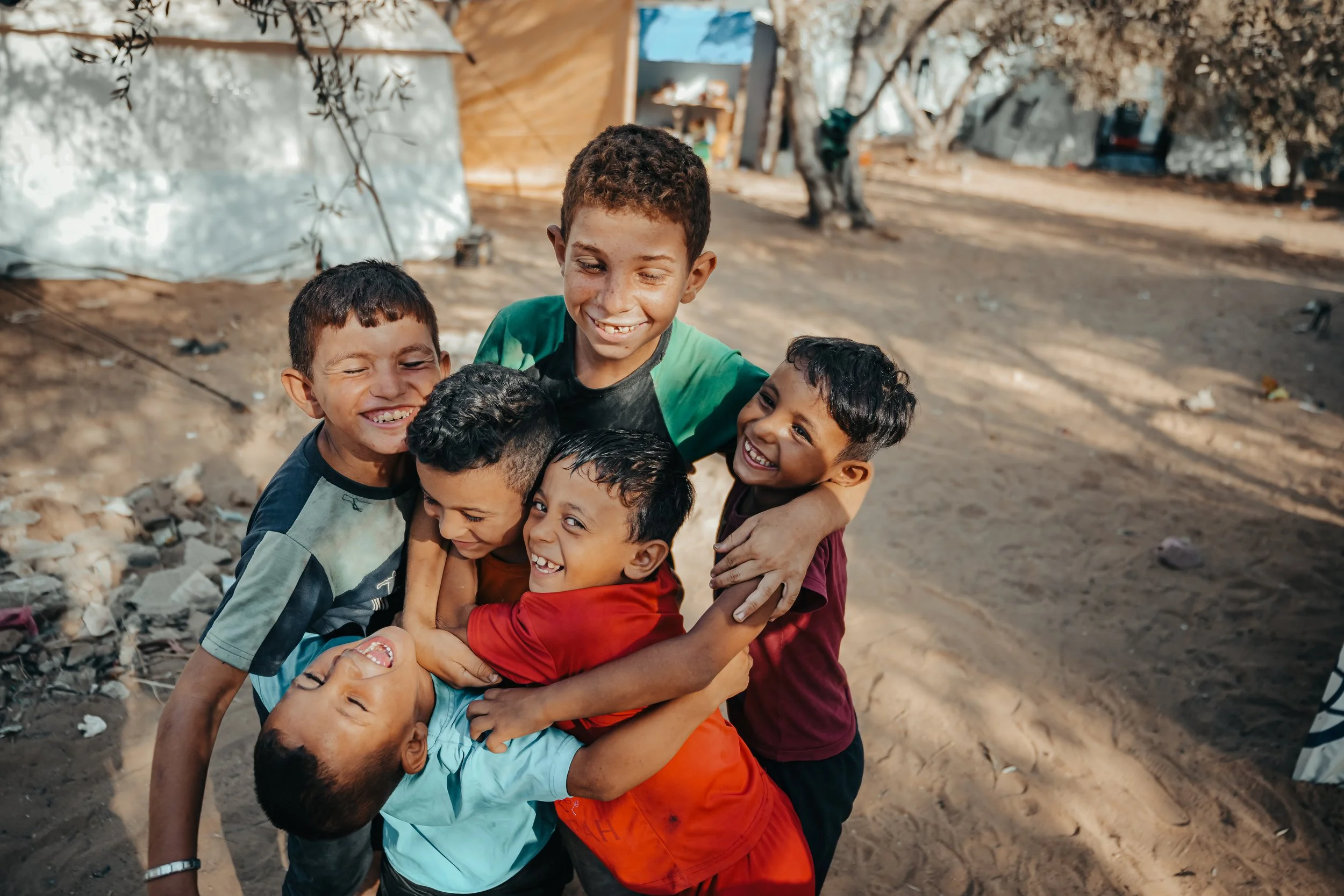 A group of Palestinian children hugging each other and smiling outdoors in a displacement tent area.