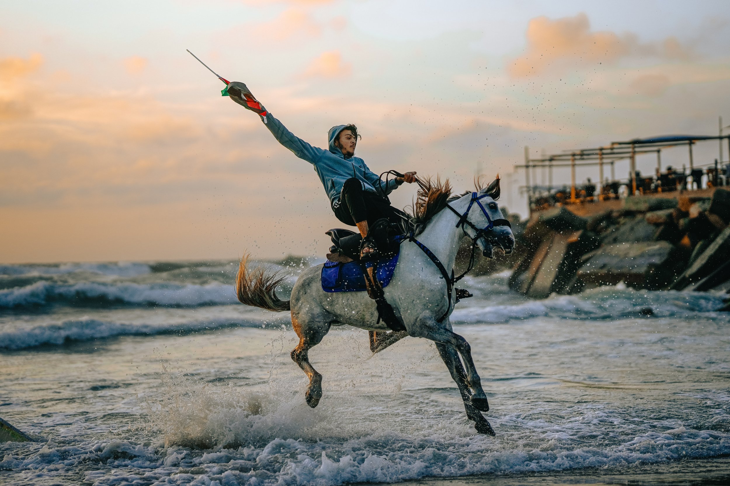 A young Palestinian man riding a horse on the beach at sunset, holding a Palestinian flag.