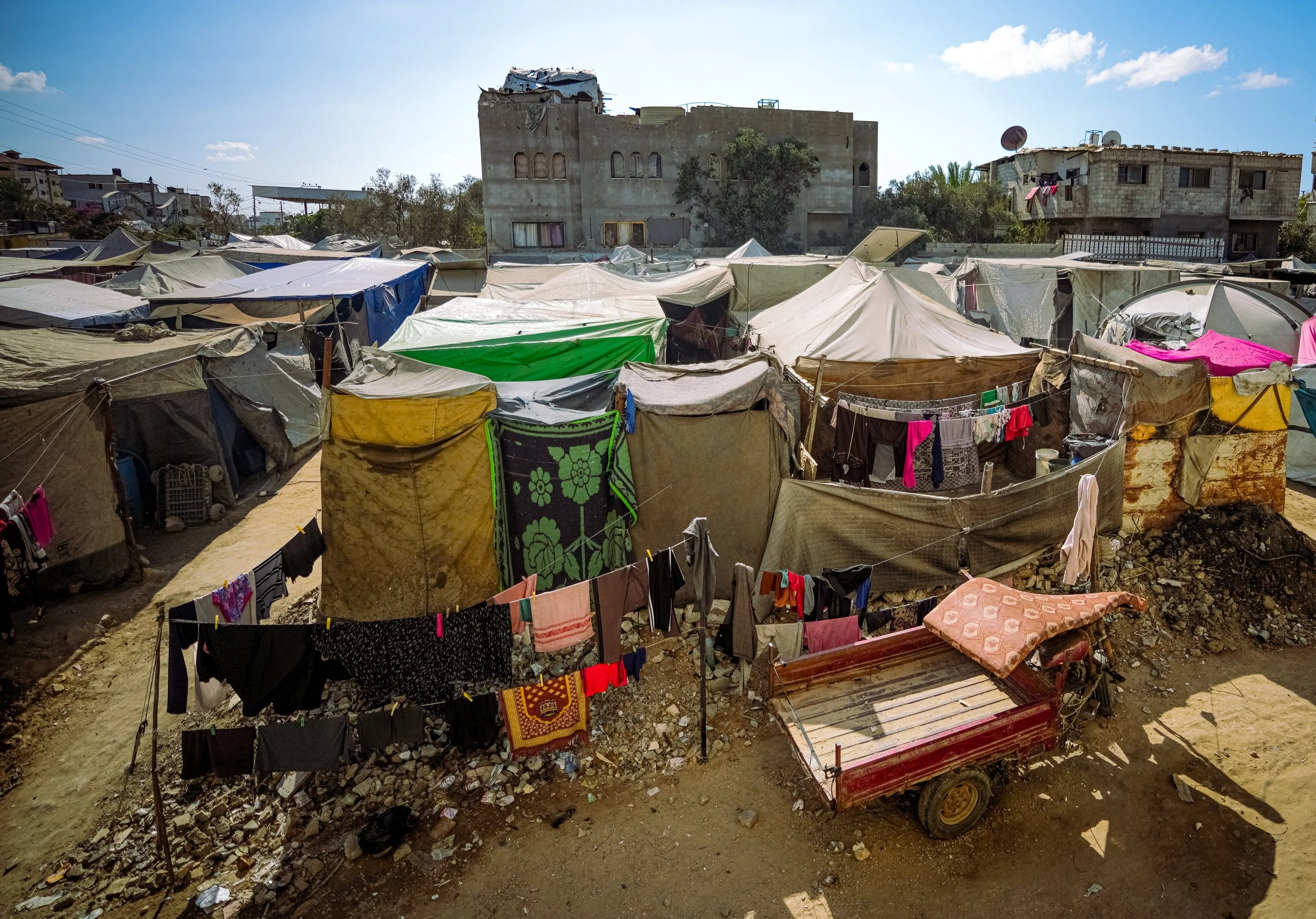 A densely packed displacement camp with makeshift tents made of fabric and plastic sheets, some clothes hanging to dry, in front of weathered buildings.