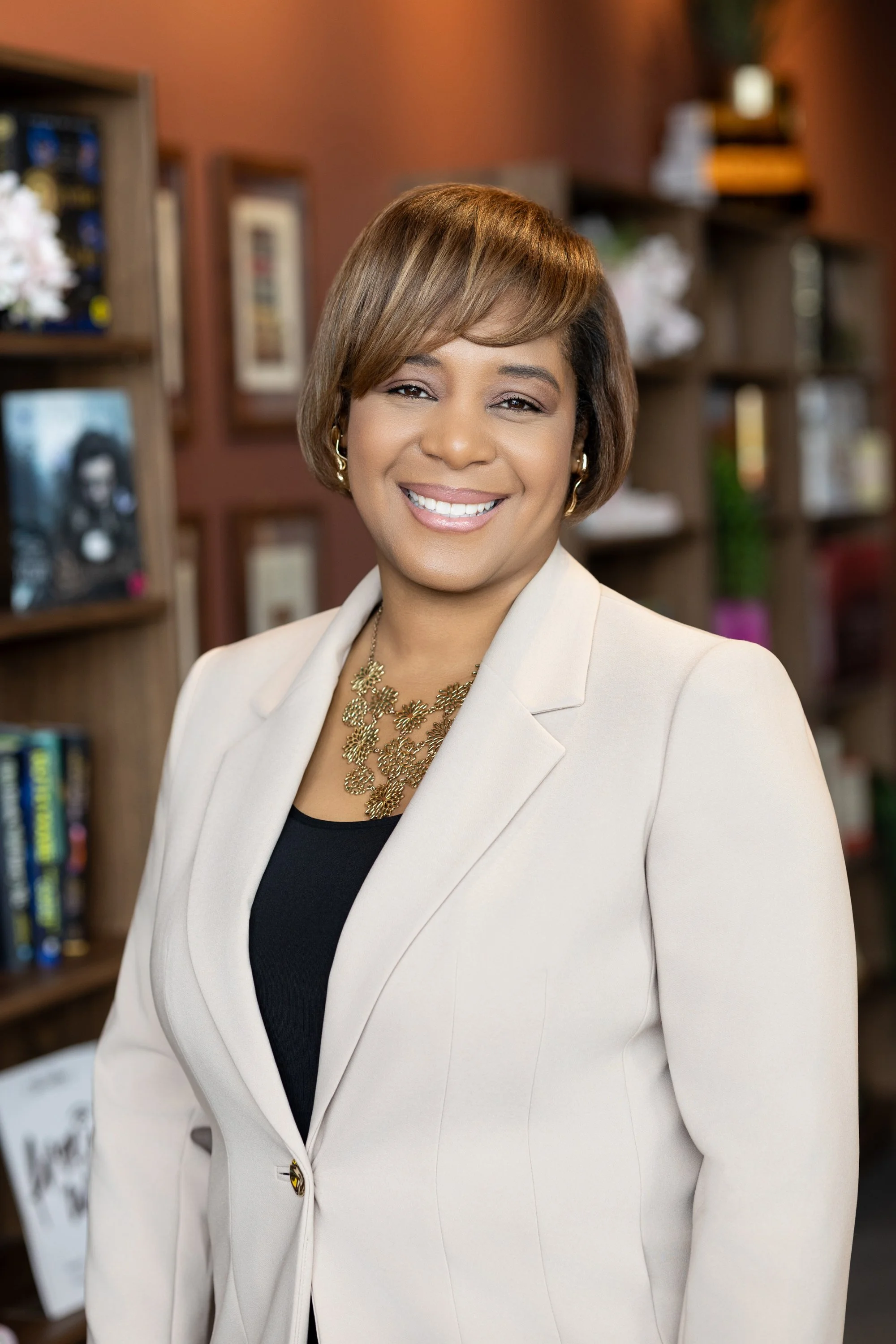 A woman with short brown hair, wearing a cream blazer and black top, smiling in an indoor setting with bookshelves and framed pictures in the background.