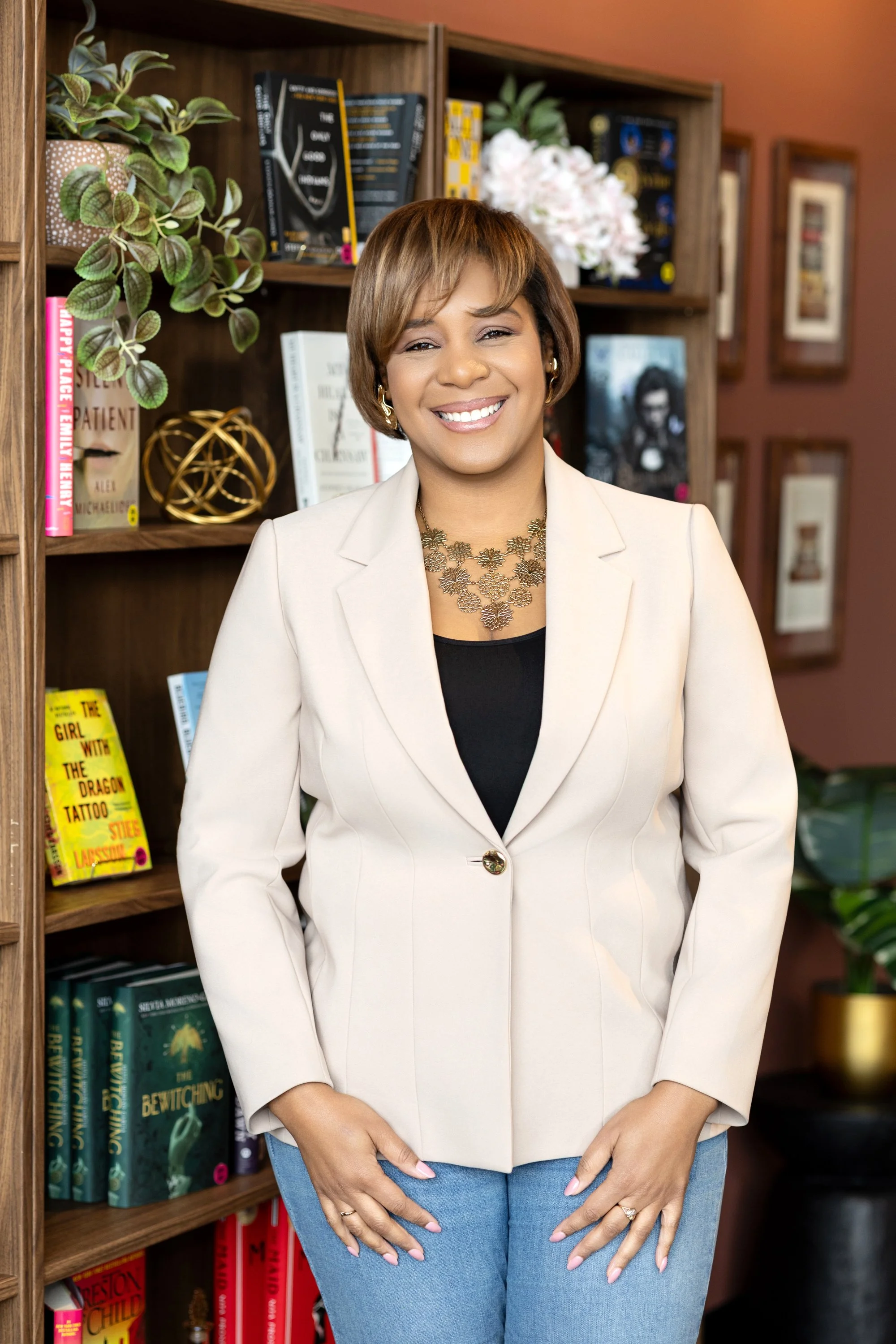 A woman with short brown hair wearing a white blazer and a black top, standing in front of a bookshelf filled with books, plants, and decorative items, smiling at the camera.