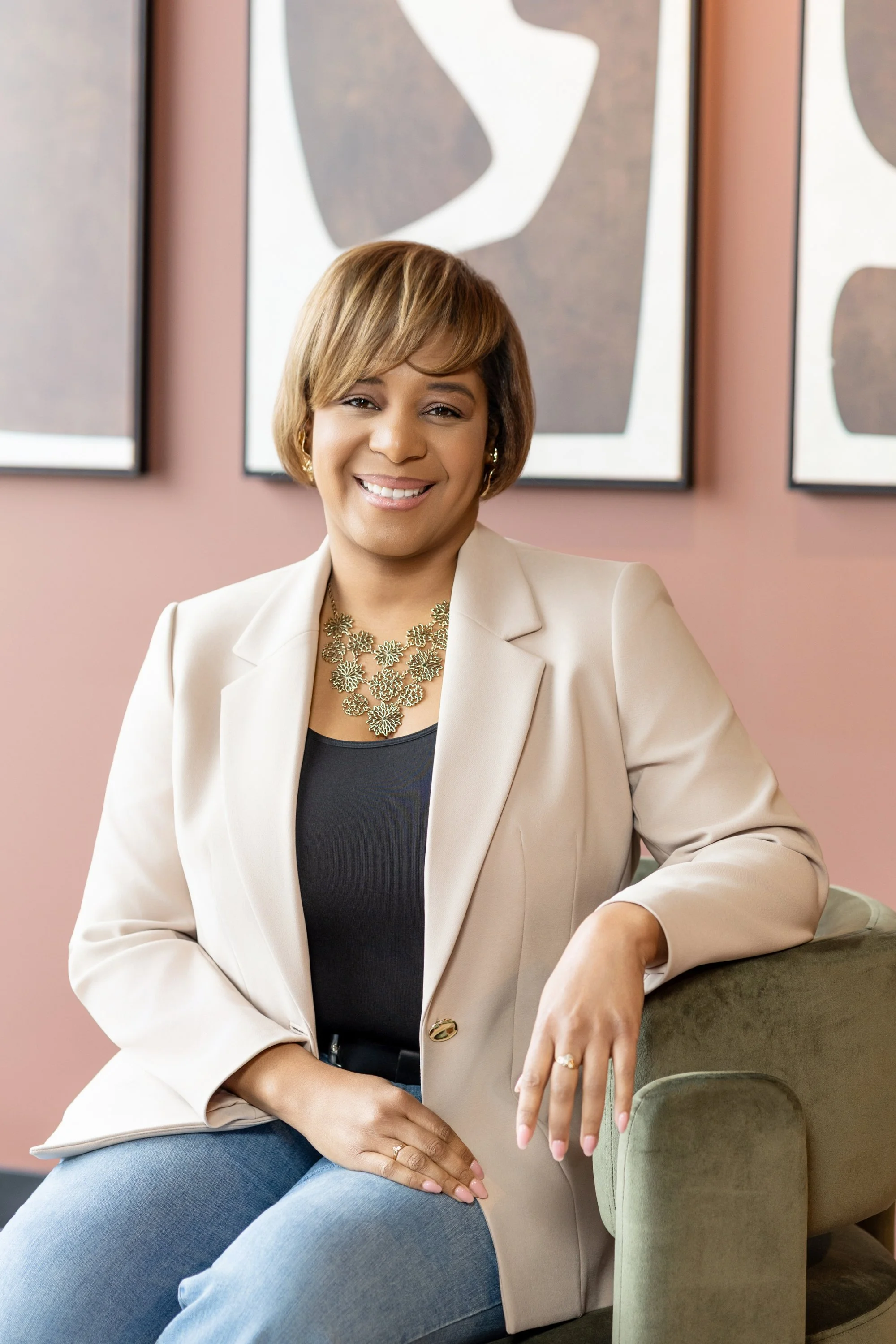 A woman smiling, sitting on a green velvet chair, wearing a beige blazer, black top, blue jeans, gold jewelry, and a ring, in front of abstract artwork on a pink wall.