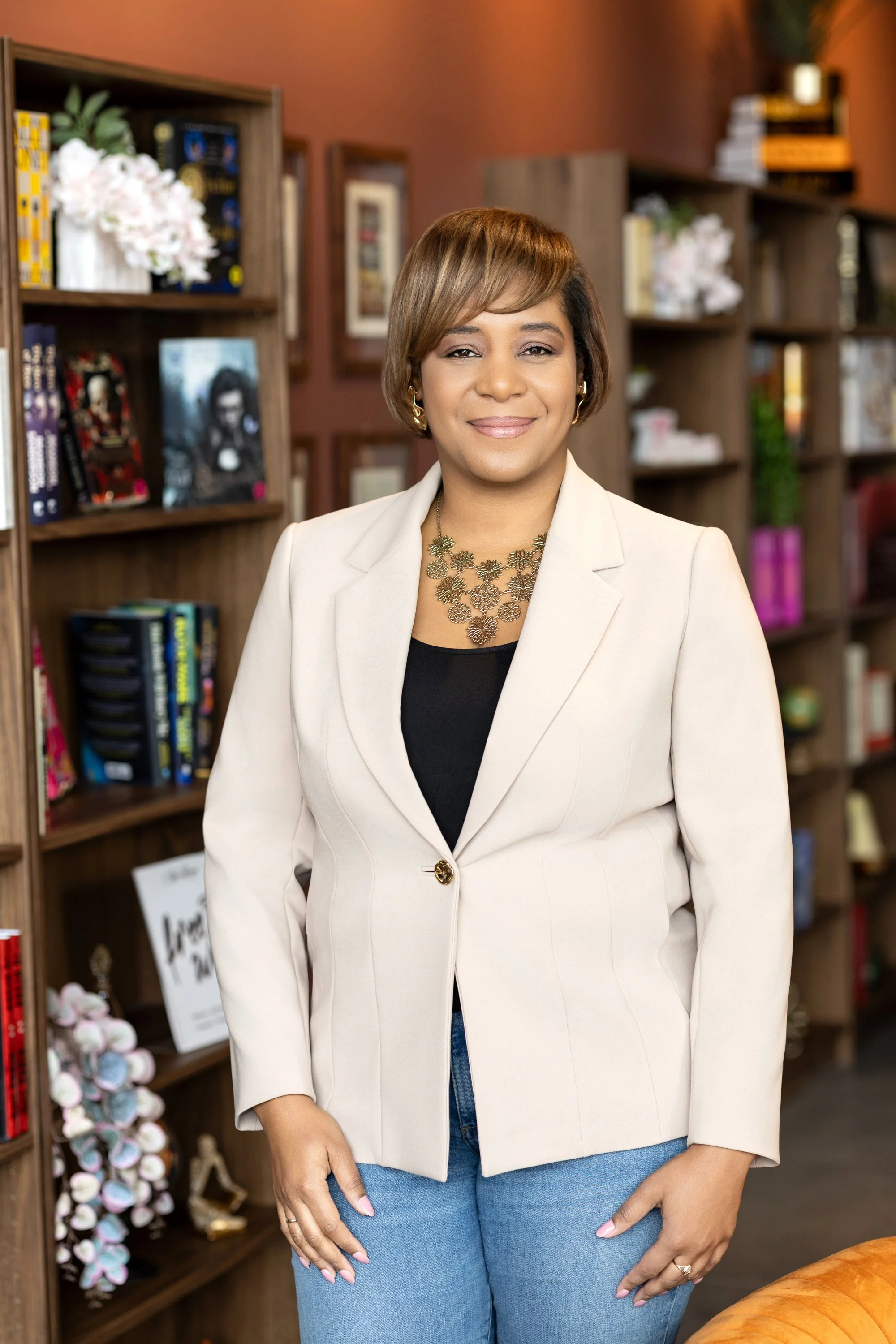A woman with shoulder-length hair, wearing a cream blazer over a black top, standing in front of a wooden bookshelf filled with books and decorative items.