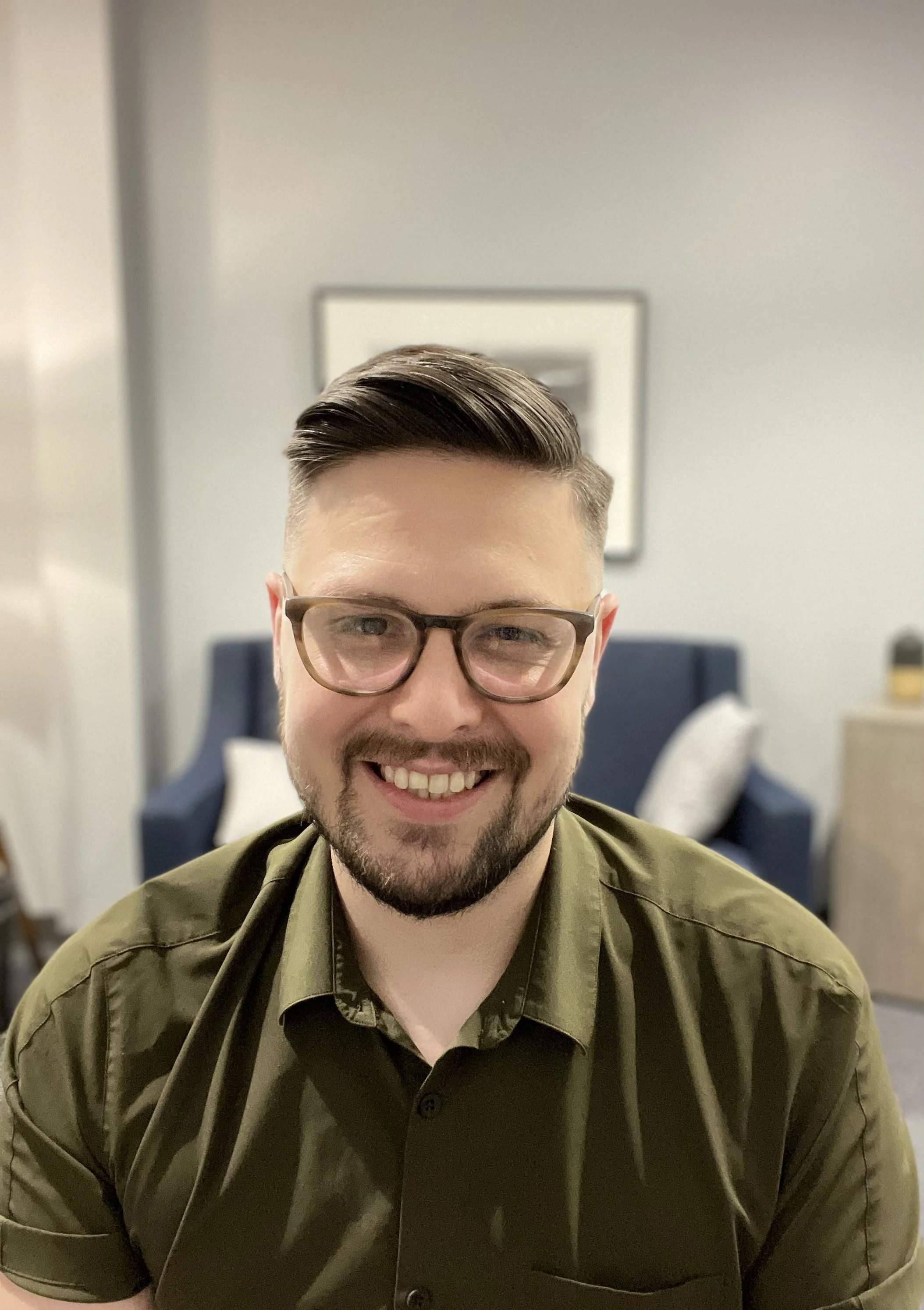 Close-up of Josh Bryant with glasses and a beard, wearing a green shirt, sitting in a room with a framed picture on the wall and a blue chair in the background.