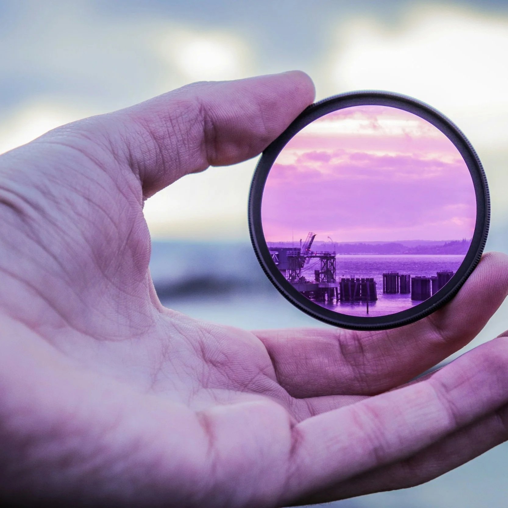 Hand holding a circular lens over a sunset by the water, reflecting buildings and clouds.