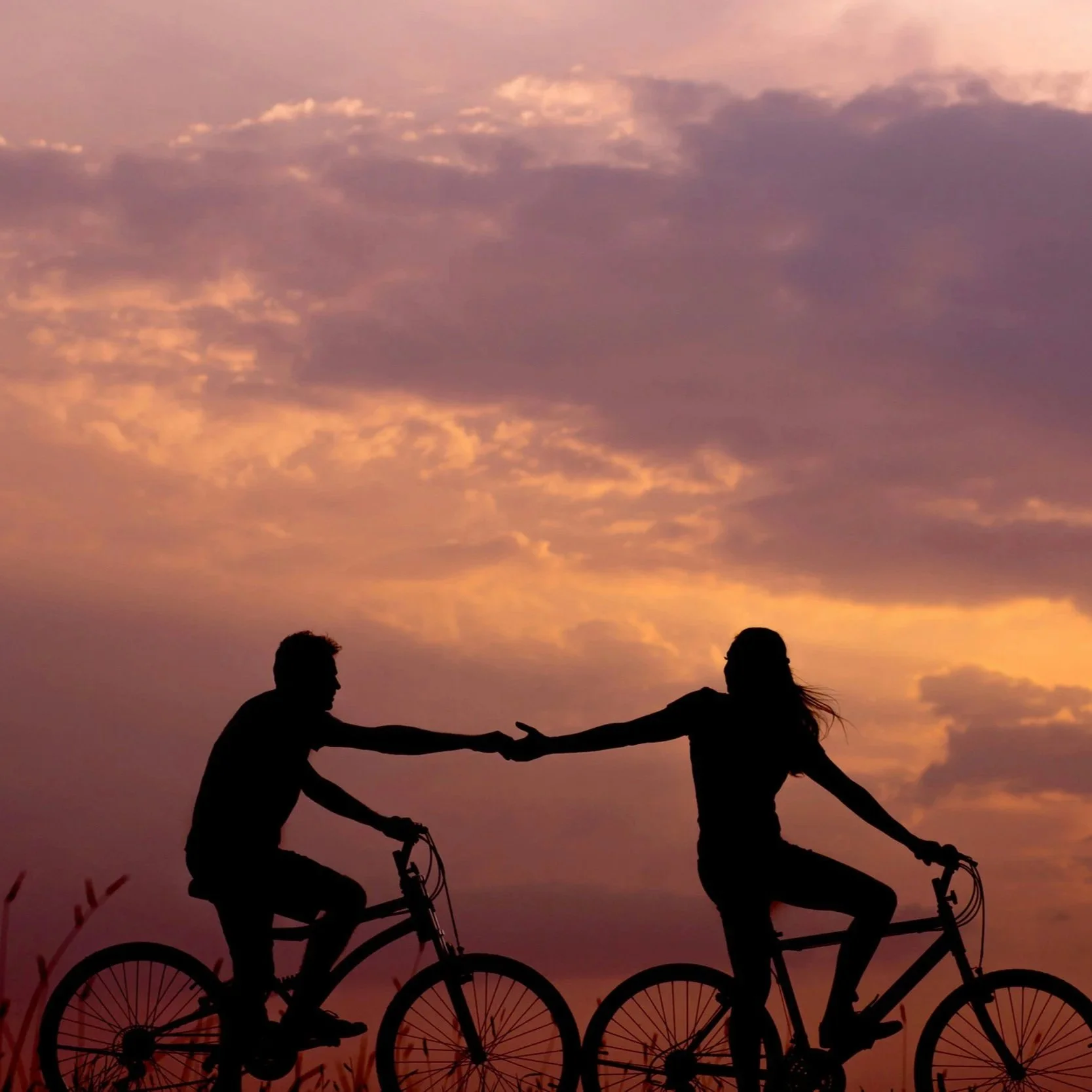 Silhouettes of two people riding bikes and holding hands against a colorful sunset sky with clouds.