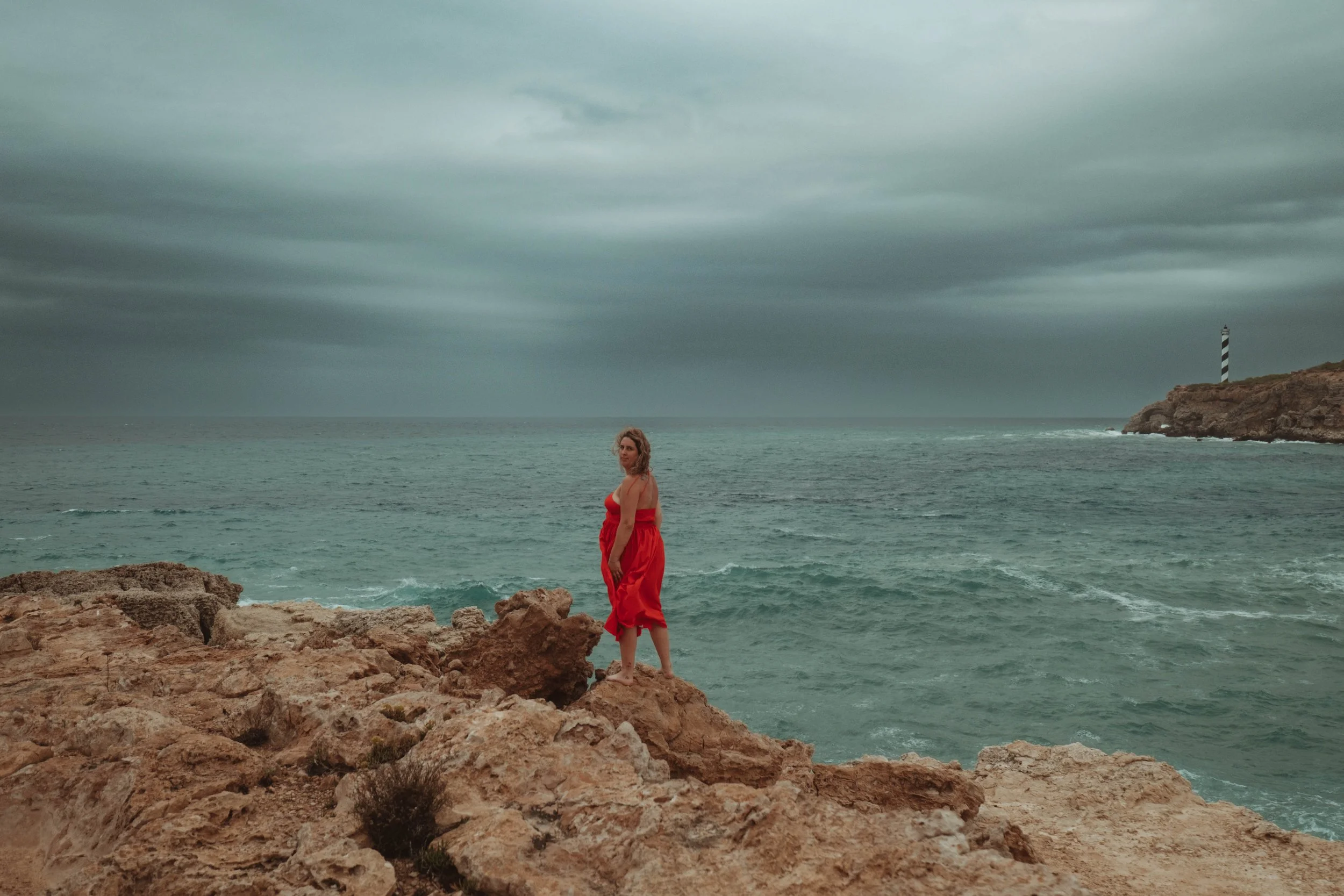 Woman in a red dress standing on rocky shore overlooking the ocean with a lighthouse in the distance under a cloudy sky.