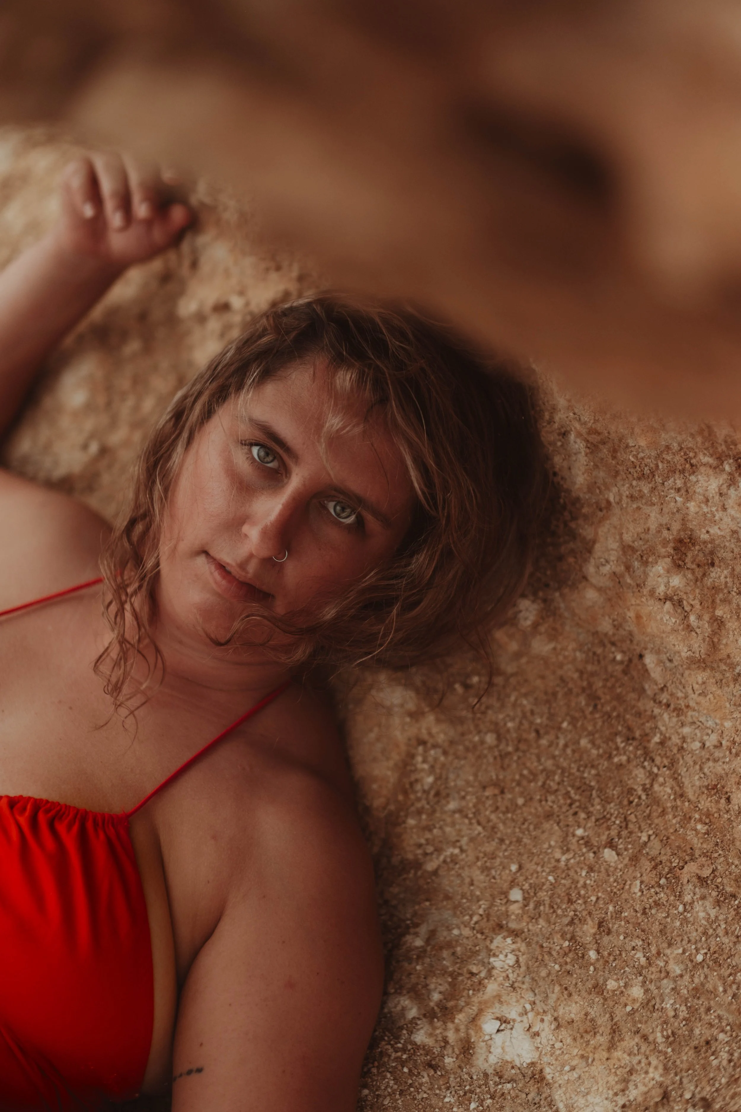 A woman with wavy, shoulder-length hair, wearing a red top, lying on the ground with her head resting on the dirt, looking directly at the camera.