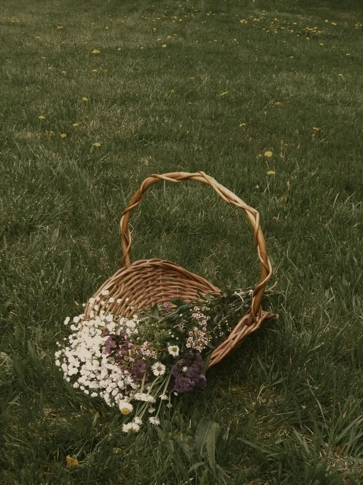 A wicker basket lying on a grassy field with small flowers spilling out of it.