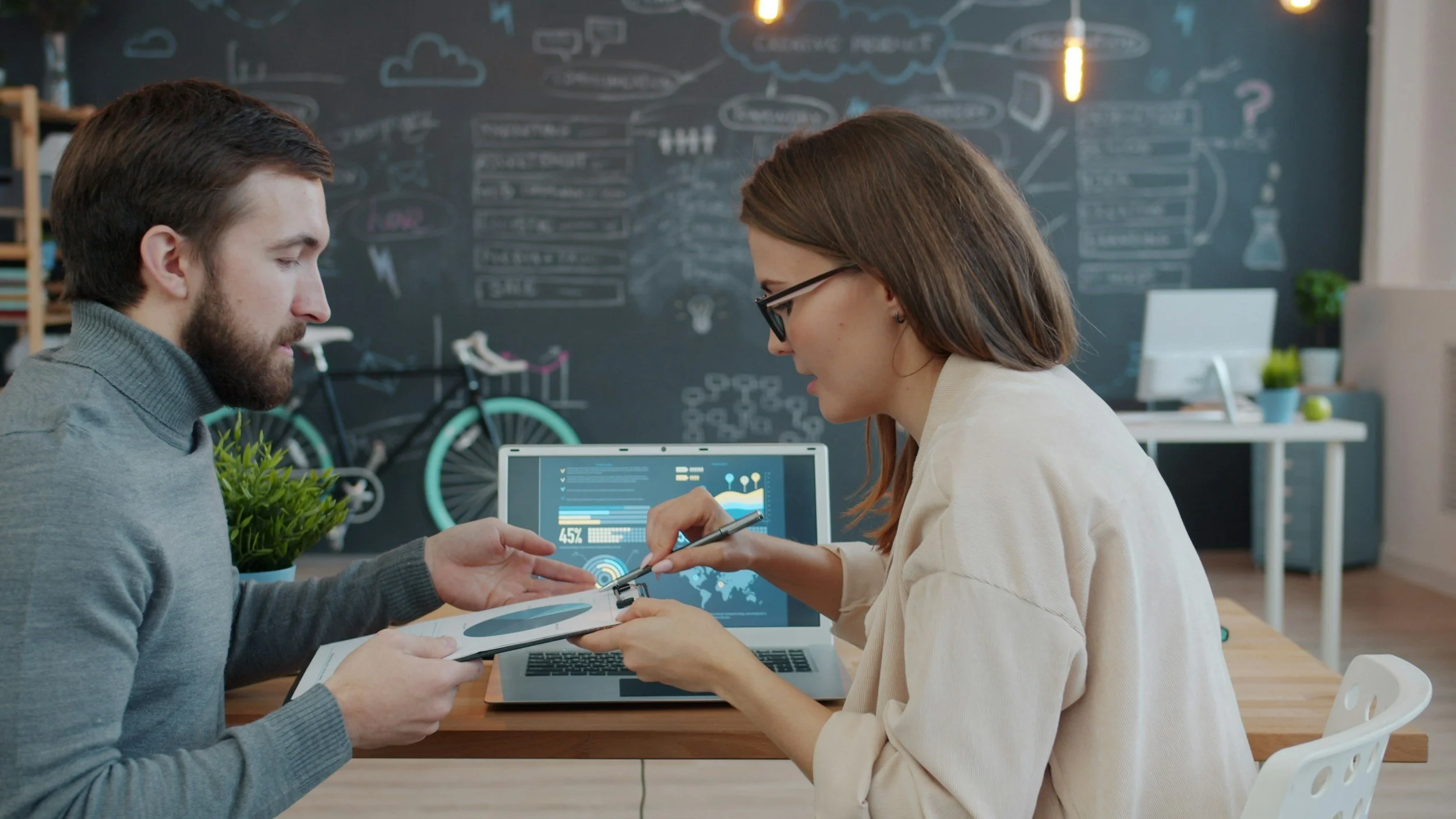 Two young professionals working together at a desk in a modern workspace with a blackboard filled with diagrams and notes in the background, and a bicycle leaning against the wall.