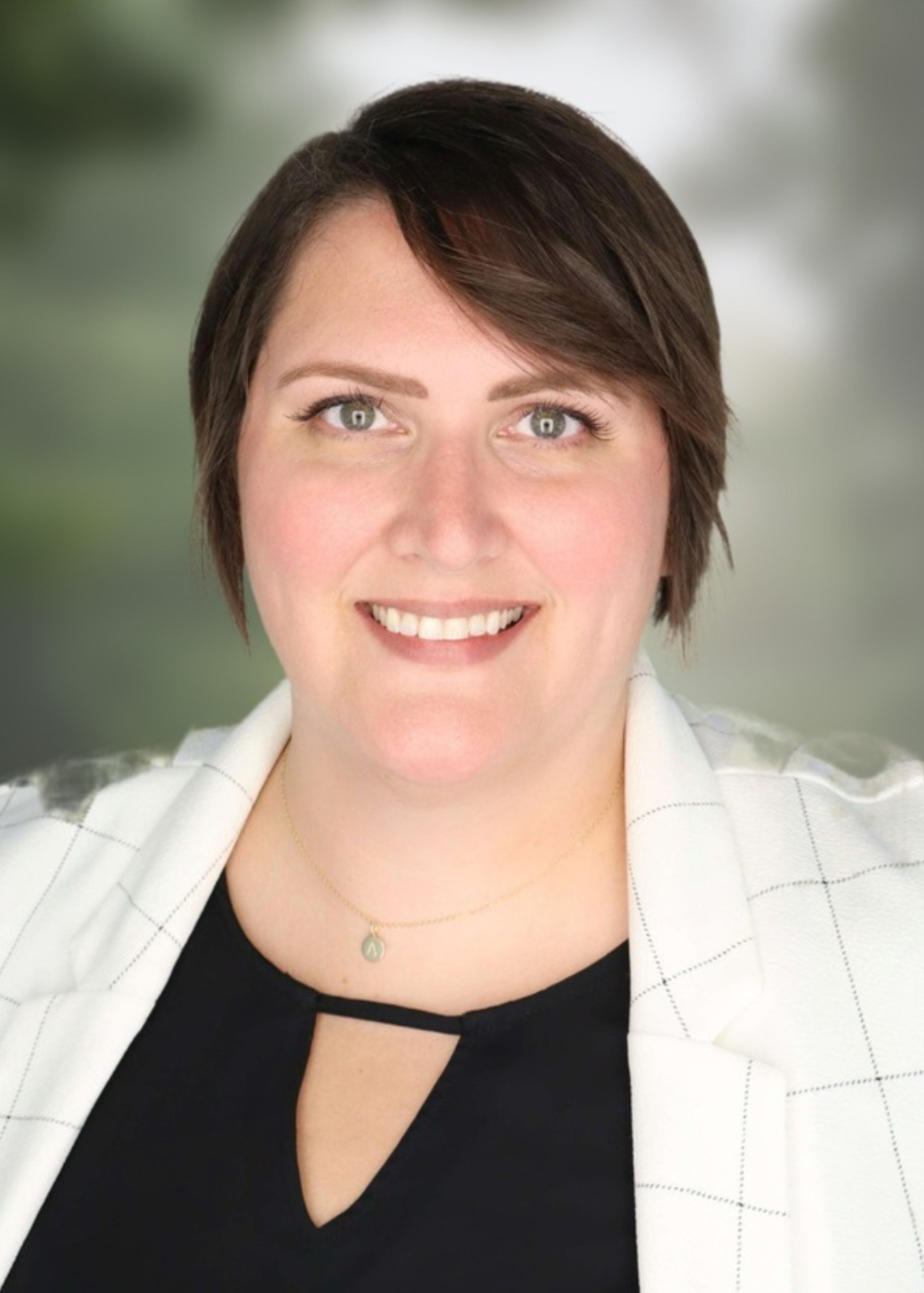 Portrait of a woman with short dark brown hair, smiling, wearing a black top, a white blazer, and a delicate necklace, with a blurred green outdoor background.