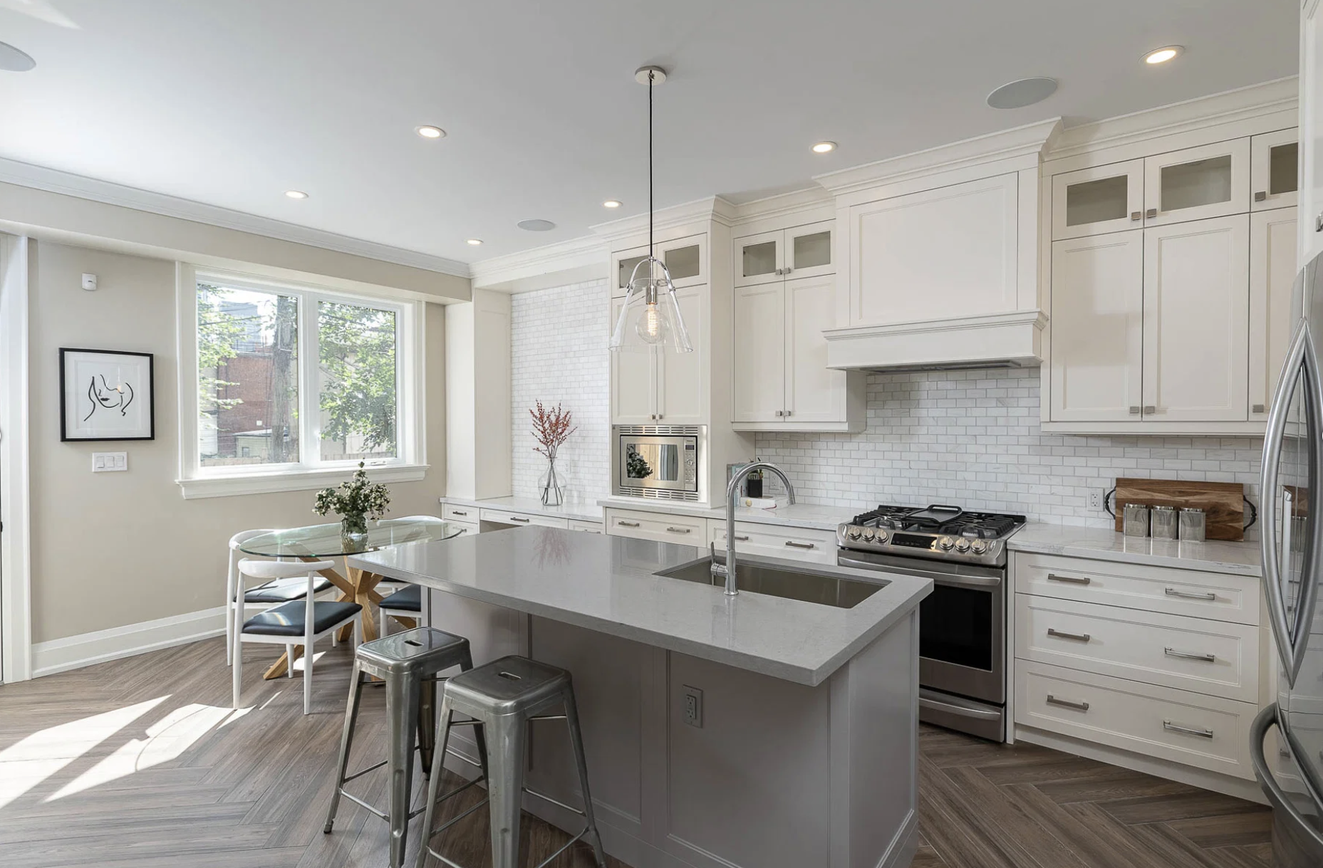 Modern white kitchen with large window, white cabinets, stainless steel appliances, kitchen island with sink, and a small dining area with a glass table and chairs.