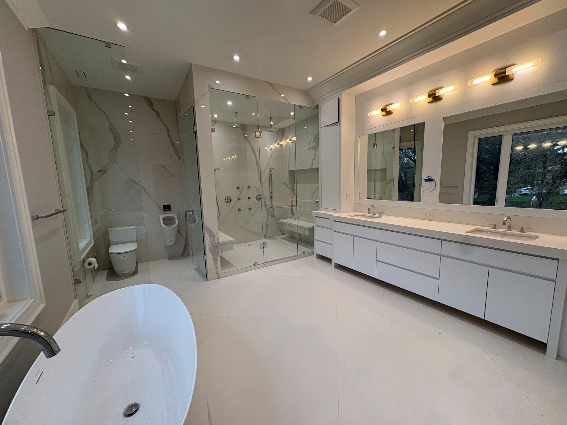 Modern bathroom with white marble floors and walls, large mirror above a white vanity with double sinks, a glass-enclosed shower with marble walls, a toilet, and a window showing outdoor trees.