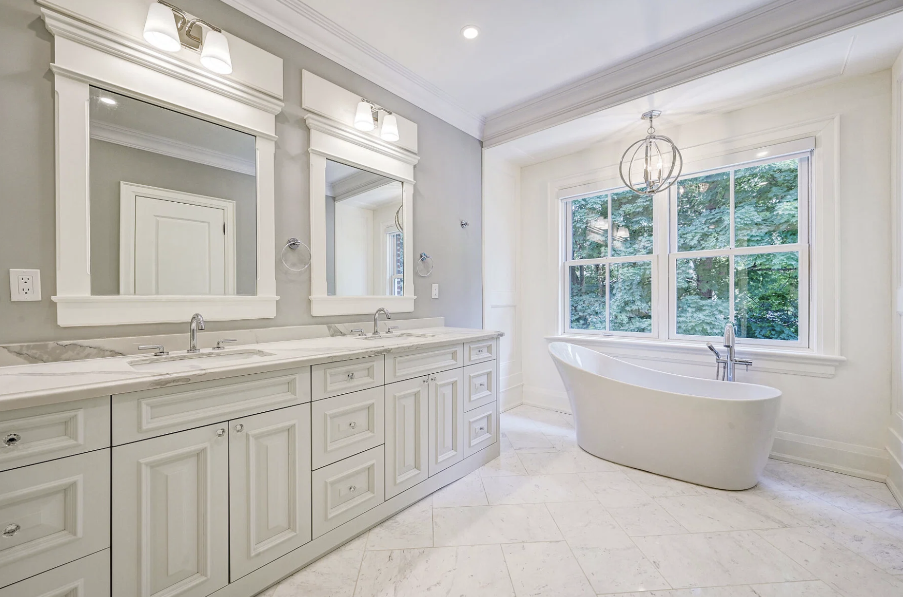 Bright modern bathroom with a double vanity, white cabinets, marble countertop, two mirrors, and two light fixtures. There is a bathtub by a large window with greenery outside.
