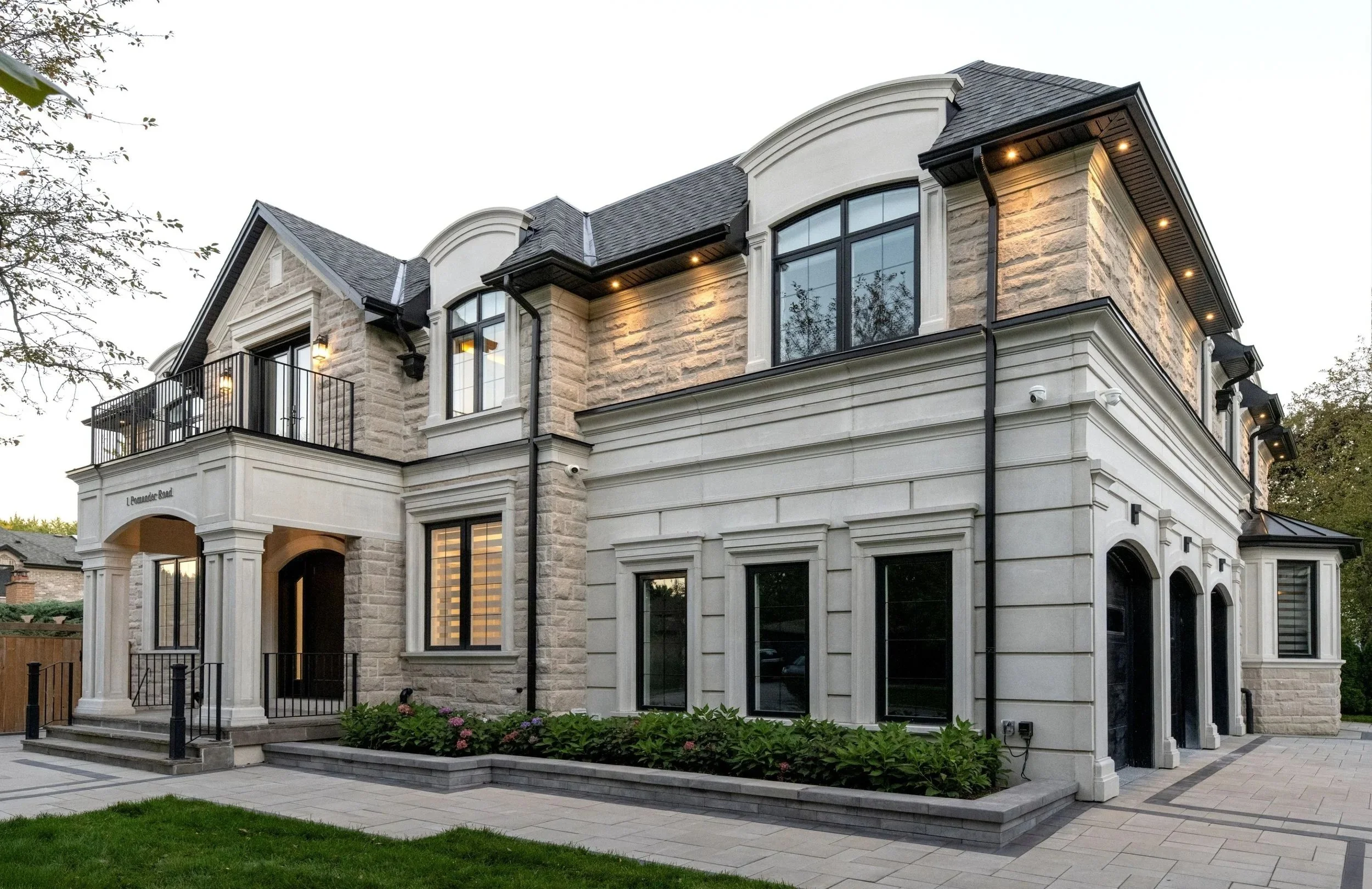 A large, multi-story house with stone and stucco exterior, multiple windows, and a balcony with a black railing. There is a ledge with garden flowers and a paved walkway in front.