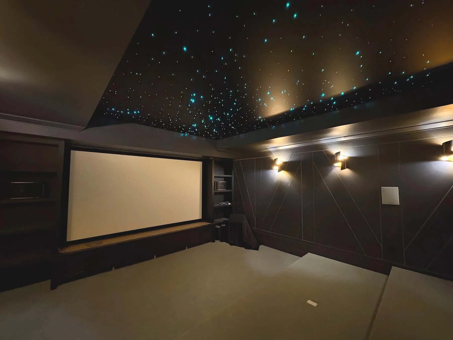 A dimly lit home theater room with a starry night ceiling, a large projector screen on the wall, and a small table with a dark cloth in front of the screen.