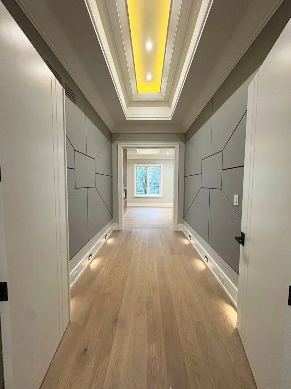 Modern hallway with gray paneled walls, recessed lighting, and hardwood flooring leading to a bright room with a window.