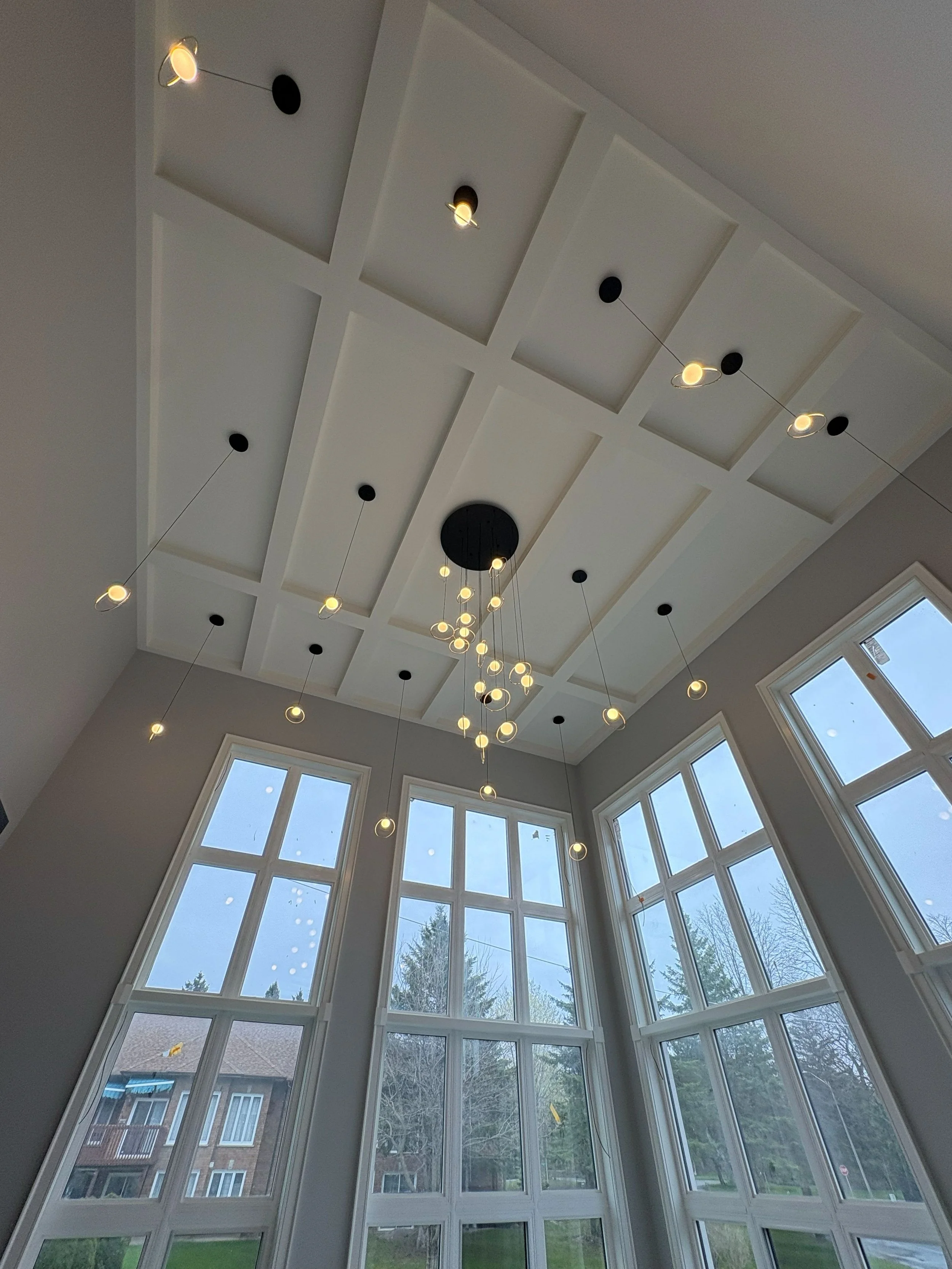 A room with a large wall of windows looking outside to trees and neighboring houses. The ceiling has a modern chandelier with multiple hanging lights and a coffered design.