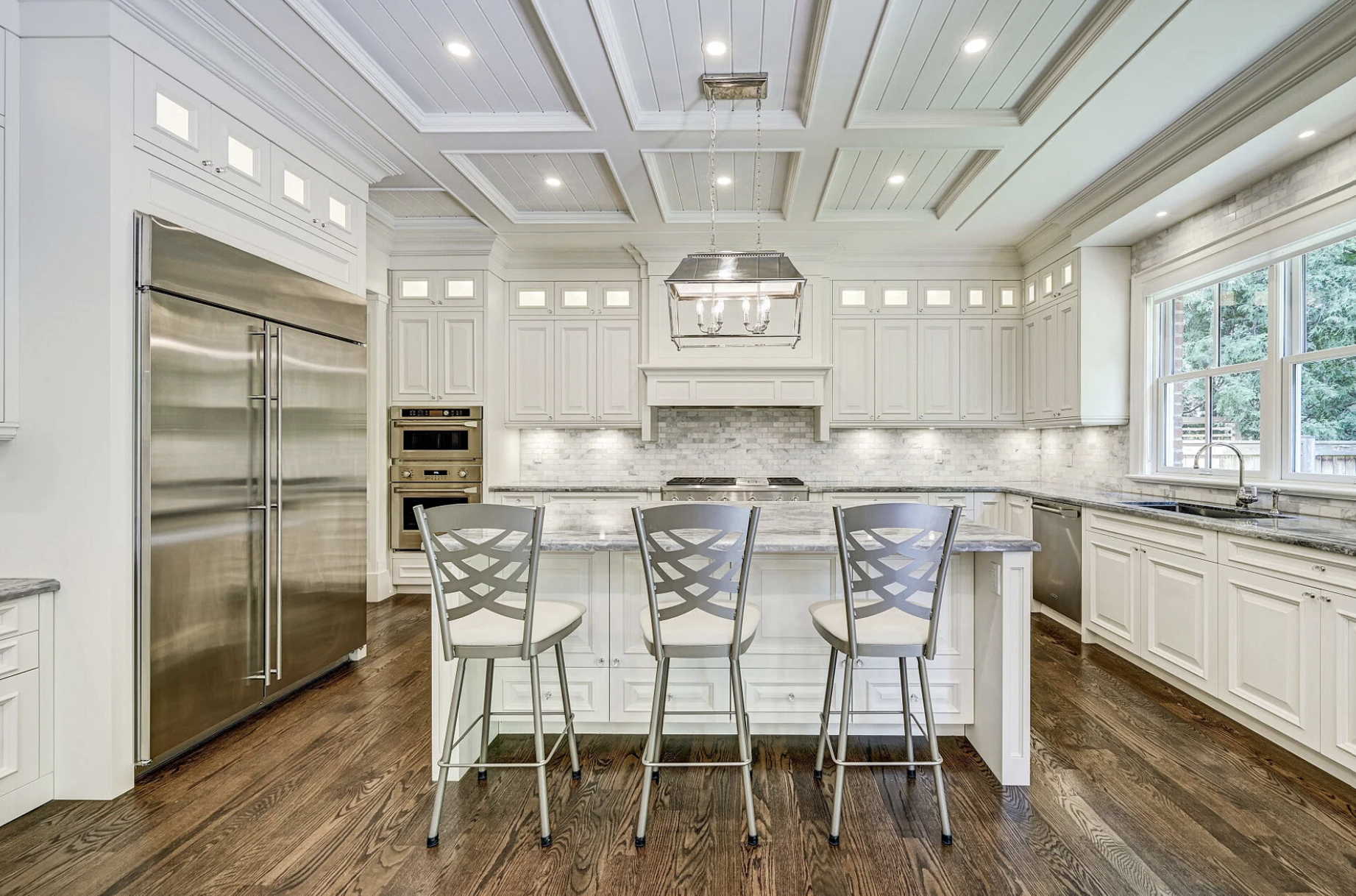 Bright, modern kitchen with white cabinetry, marble countertops, a large island with four chairs, stainless steel appliances including a double-door refrigerator, built-in oven and microwave, and a large window overlooking a backyard. Recessed lighti
