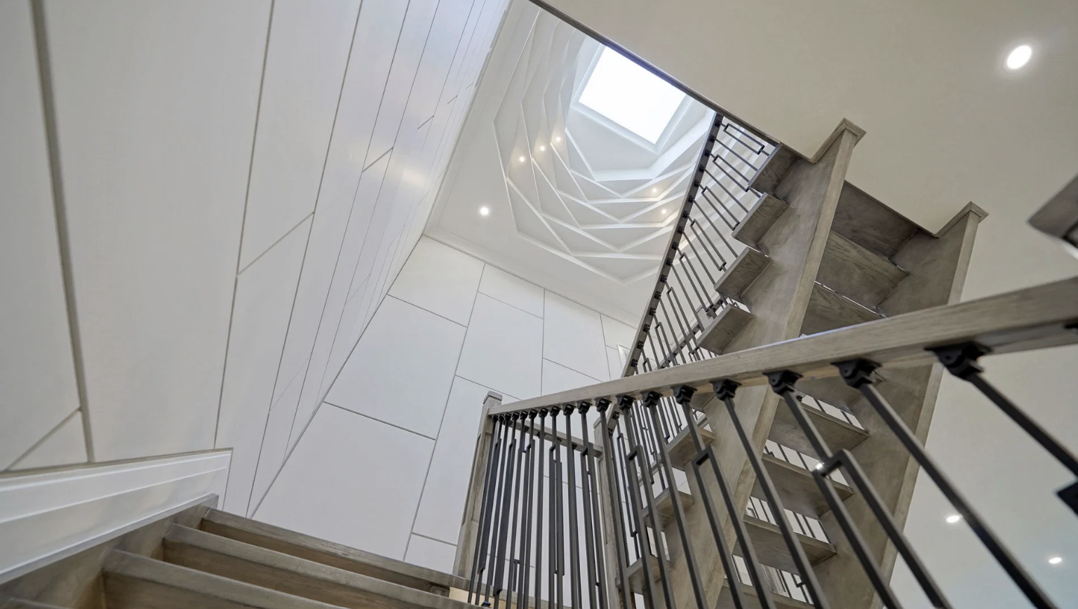 View of a staircase from below, looking up towards a skylight and spiral pattern ceiling with recessed lighting.