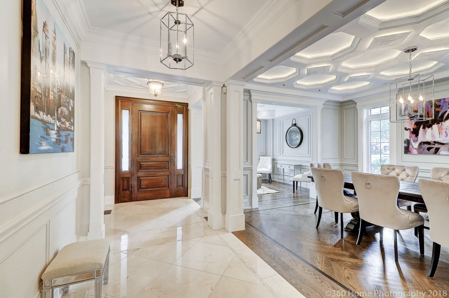 Entrance foyer with wooden front door, marble flooring, and white wainscoting; leads to an open dining area with large windows, light-colored chairs, and elegant decor.