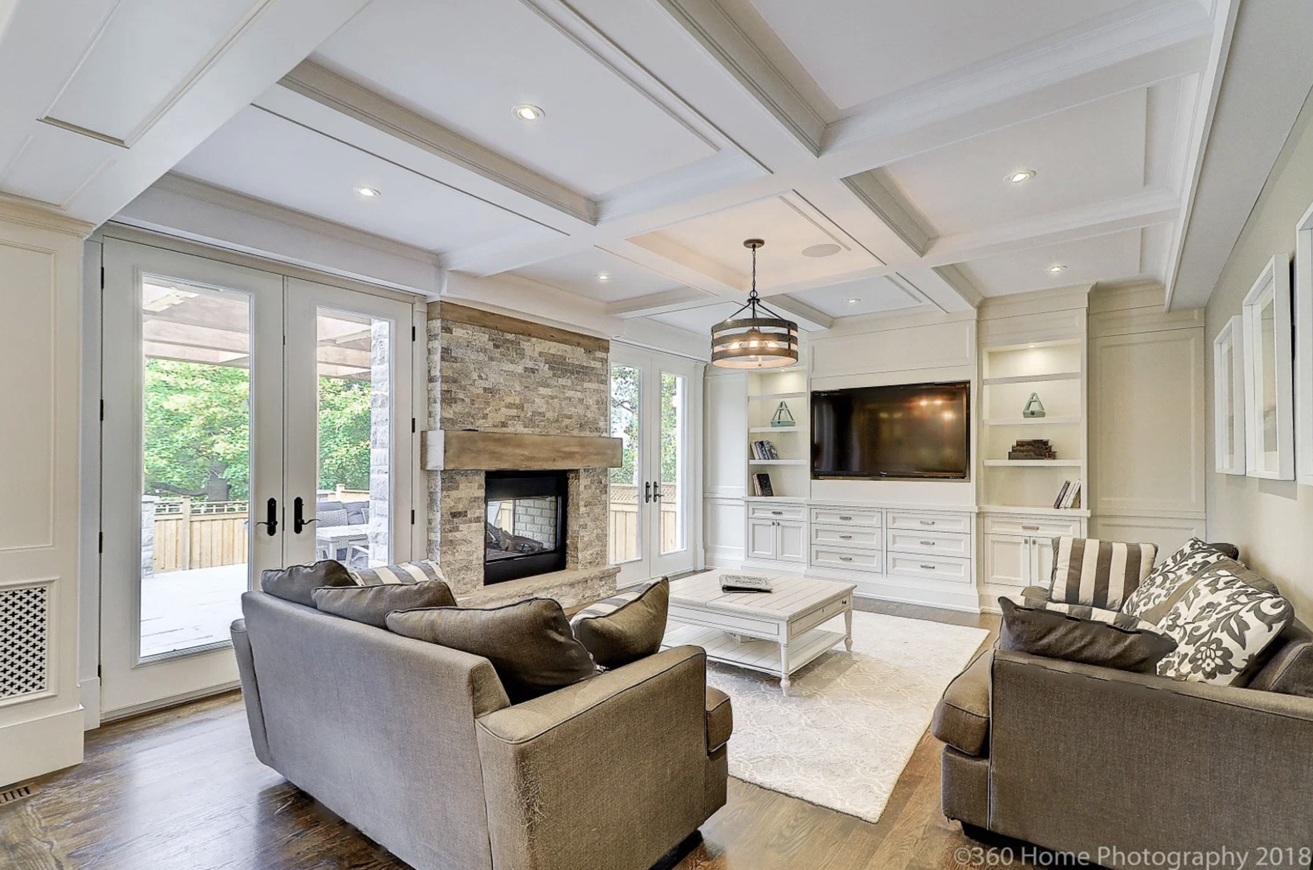 Living room with white built-in shelves, a flat-screen TV, a stone fireplace, glass doors leading outside, and brown sofas with patterned and solid cushions.