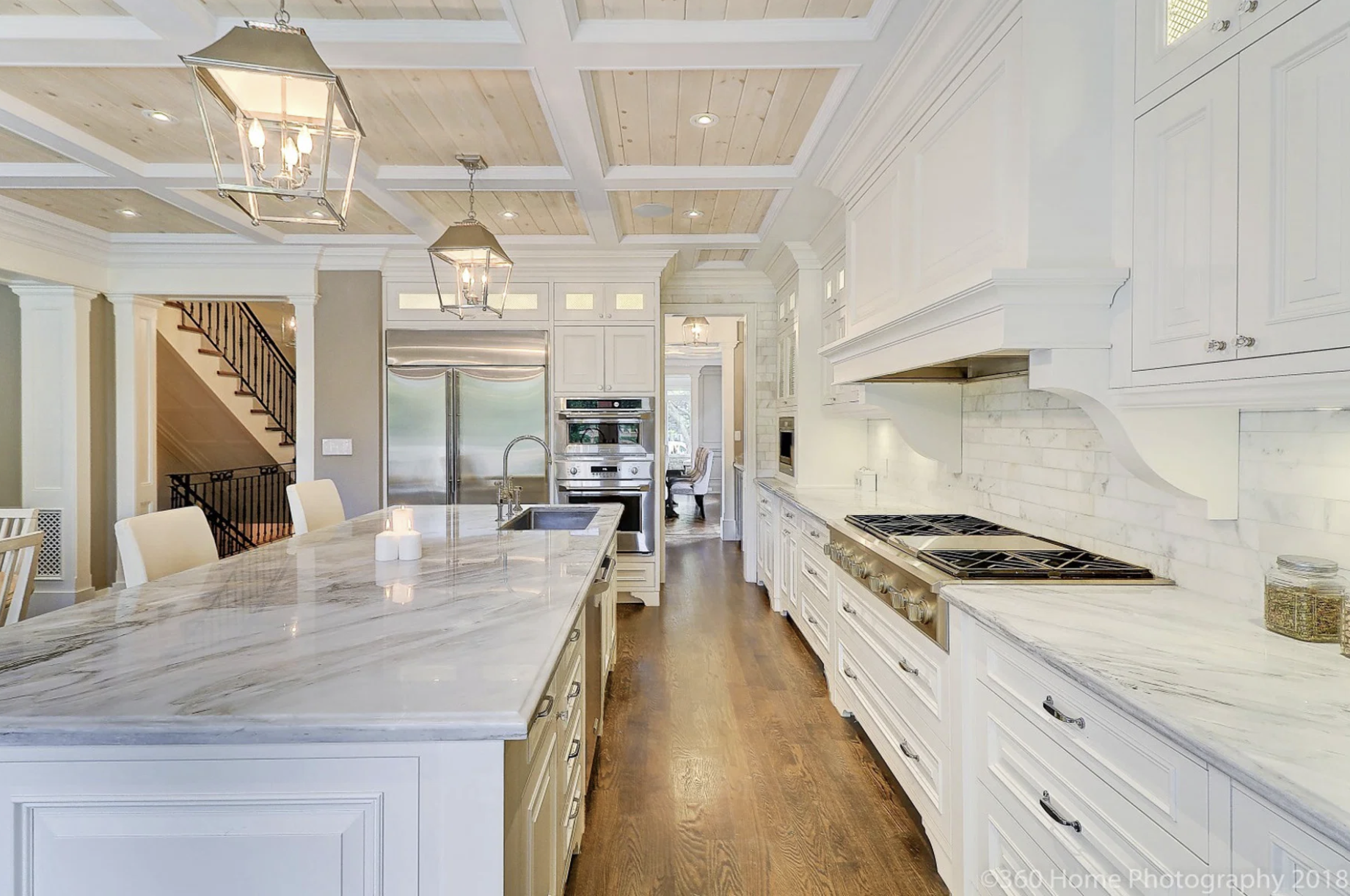 A spacious, modern kitchen with white cabinetry, marble countertops, and a large island with seating. Stainless steel appliances, including a refrigerator, oven, and microwave, are visible. The ceiling has wooden paneling with recessed lighting and h