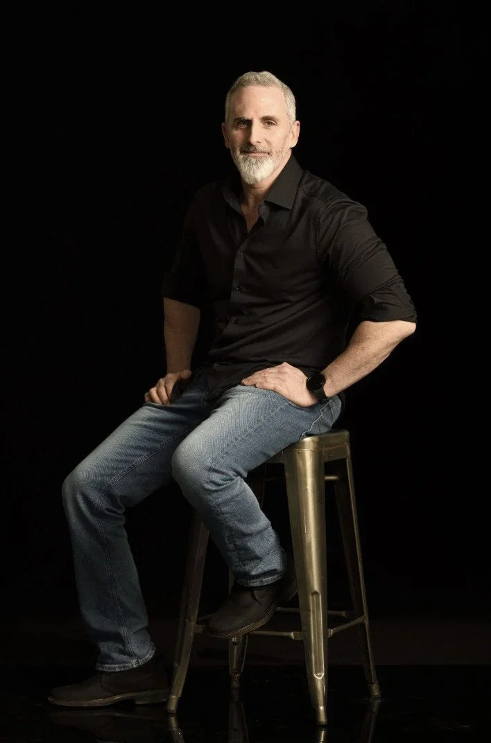 A middle-aged man with a gray beard and short gray hair sitting on a tall metal stool against a black background. He is wearing a black button-up shirt, blue jeans, and dark shoes, looking at the camera with a slight smile.