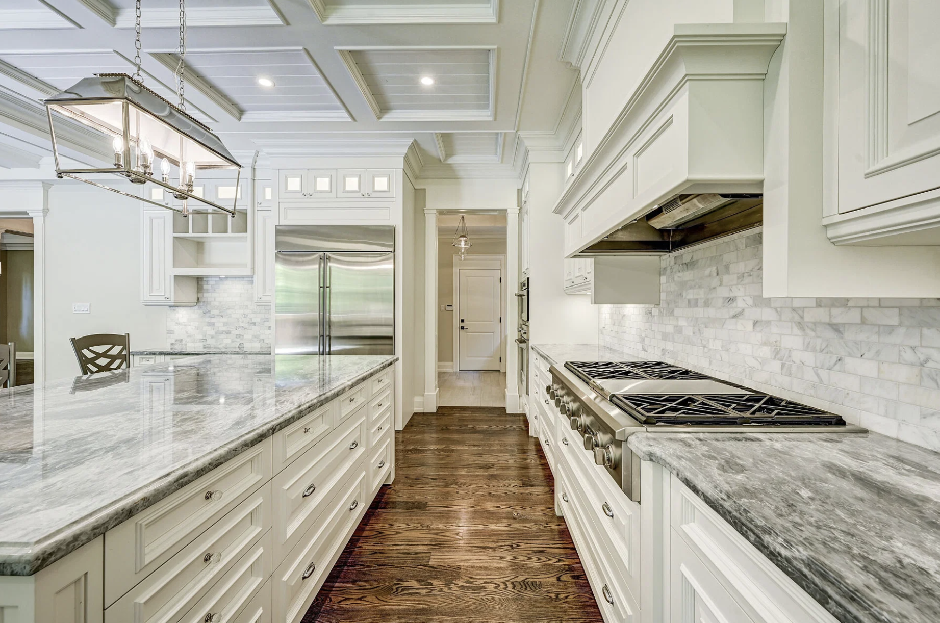 A spacious, white kitchen with marble countertops, white cabinetry, and a stainless steel refrigerator. The kitchen features a gas stove, marble tile backsplash, and dark hardwood flooring.