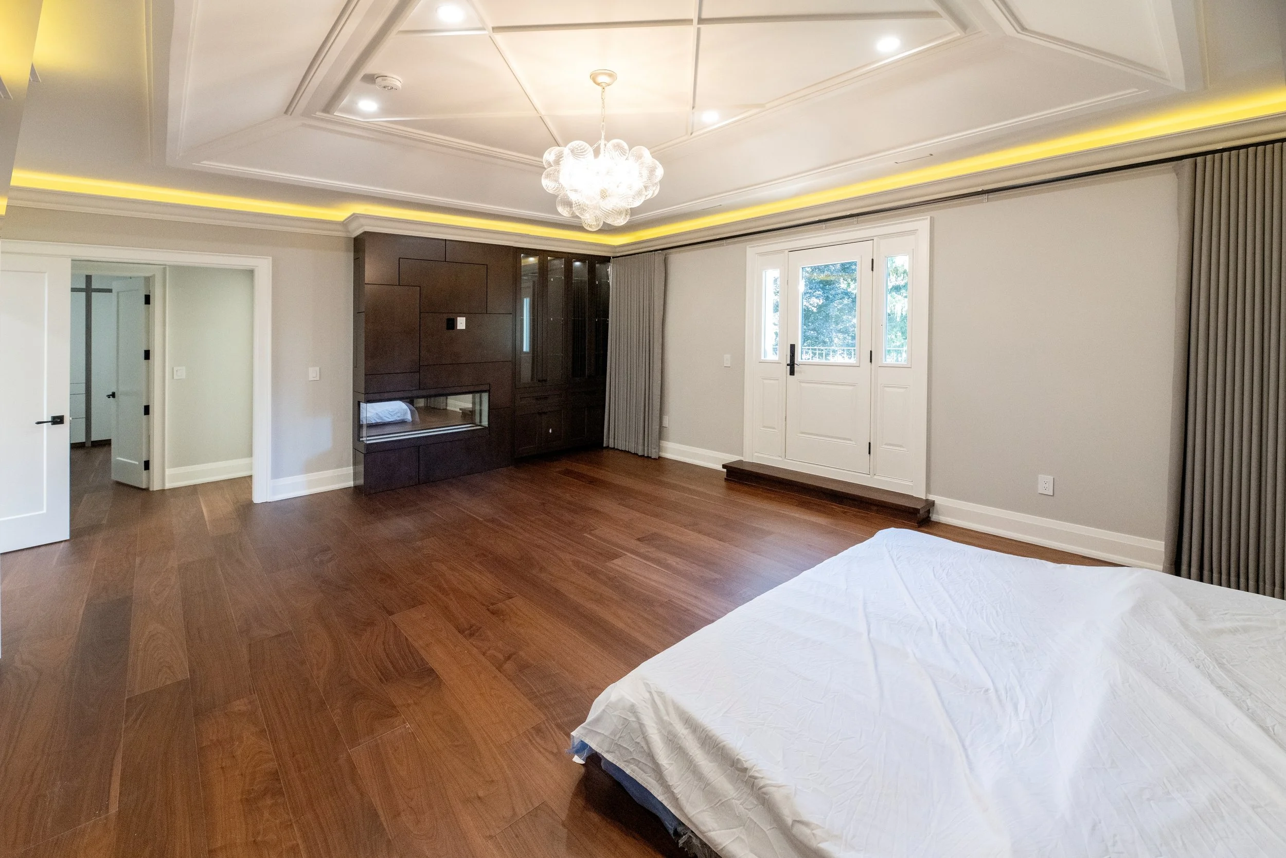 A bedroom with wooden flooring, a large window and door with curtains, a built-in dark wood cabinet, and a modern chandelier hanging from the ceiling.