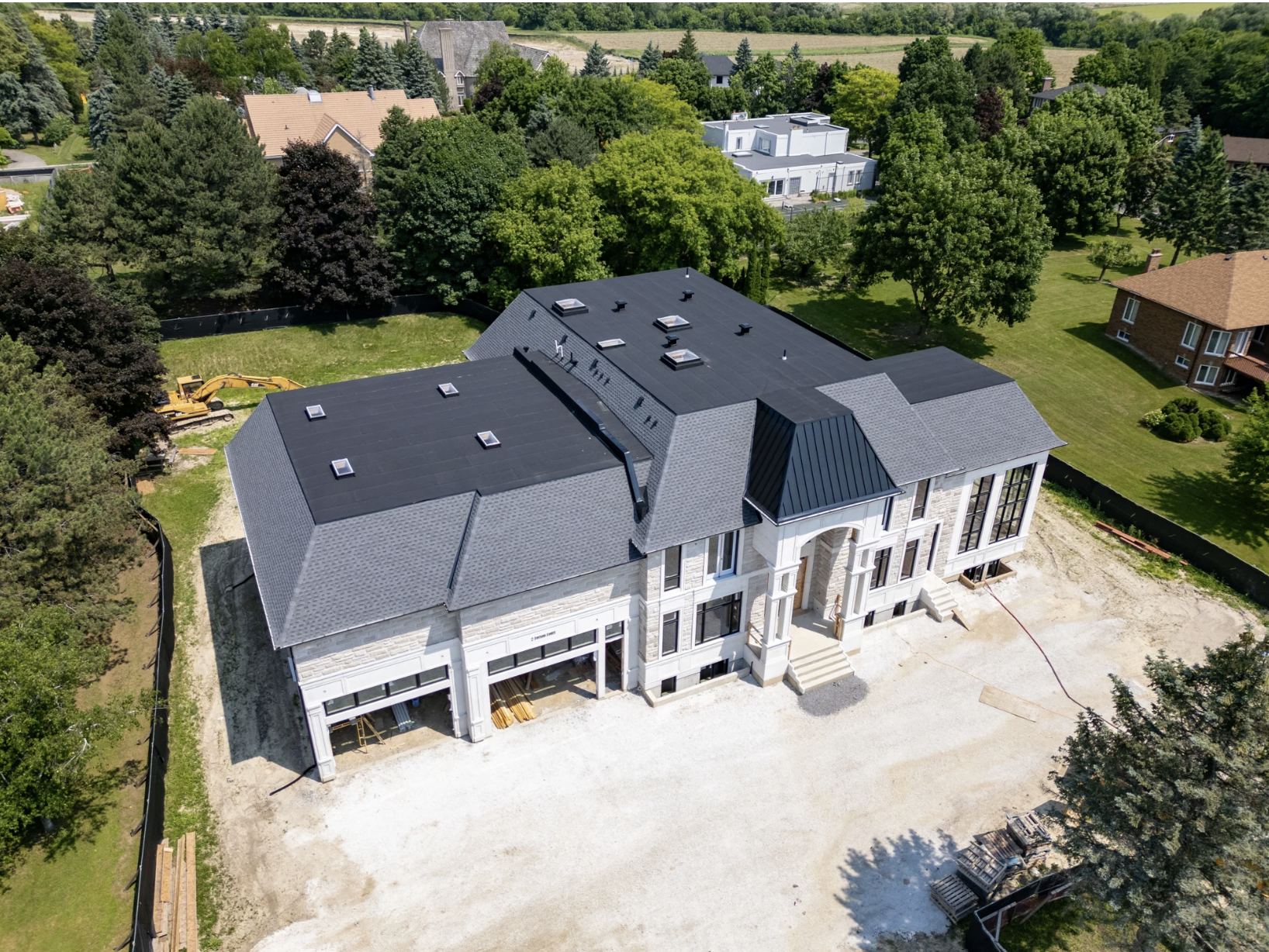 An aerial view of a large, multi-story house under construction, with a black roof and stone facade, surrounded by green trees and other residential buildings in the background.