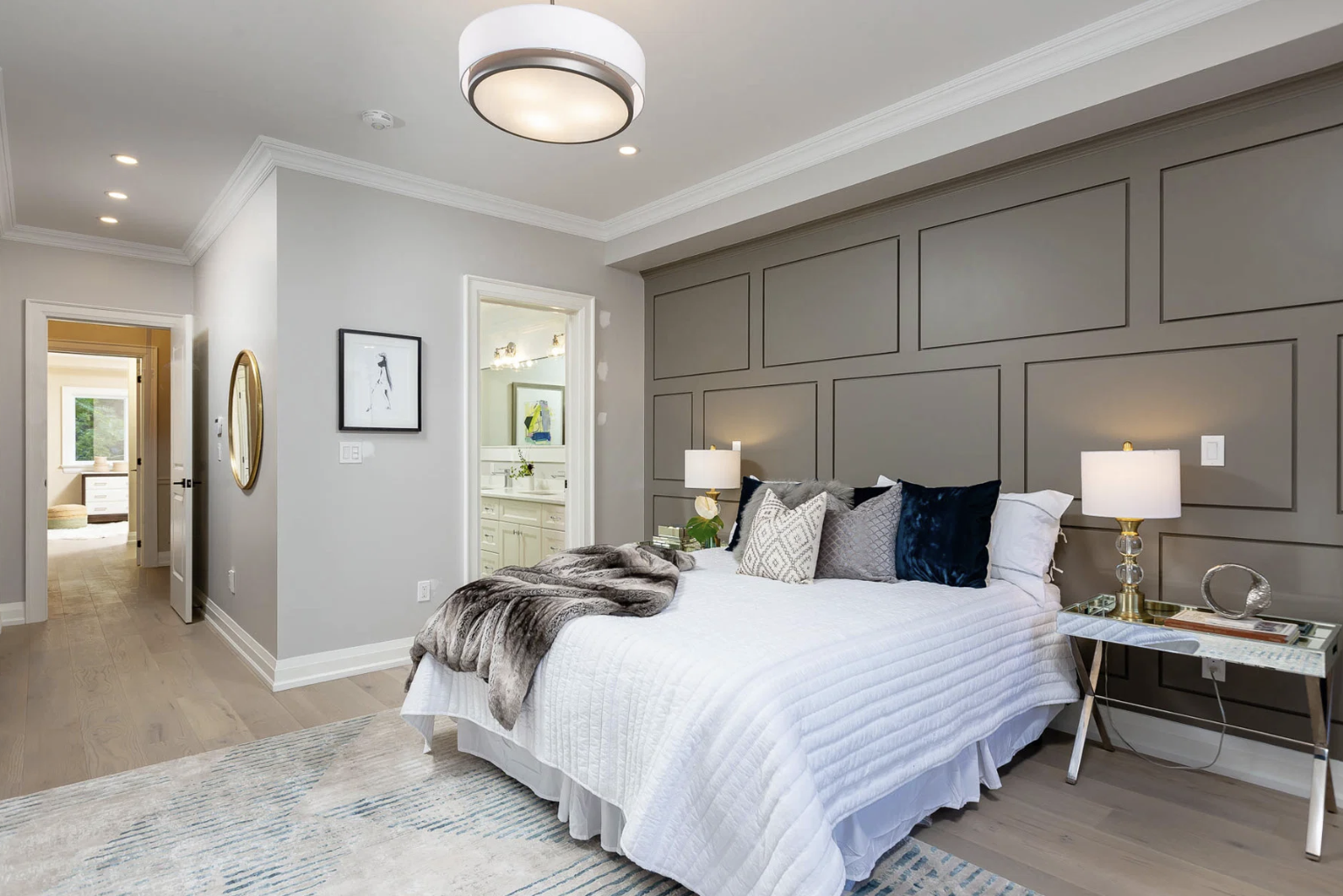 Modern bedroom with gray paneled accent wall, white bed with pillows and a faux fur throw, side table with lamp, and a doorway leading to a bathroom and another room.
