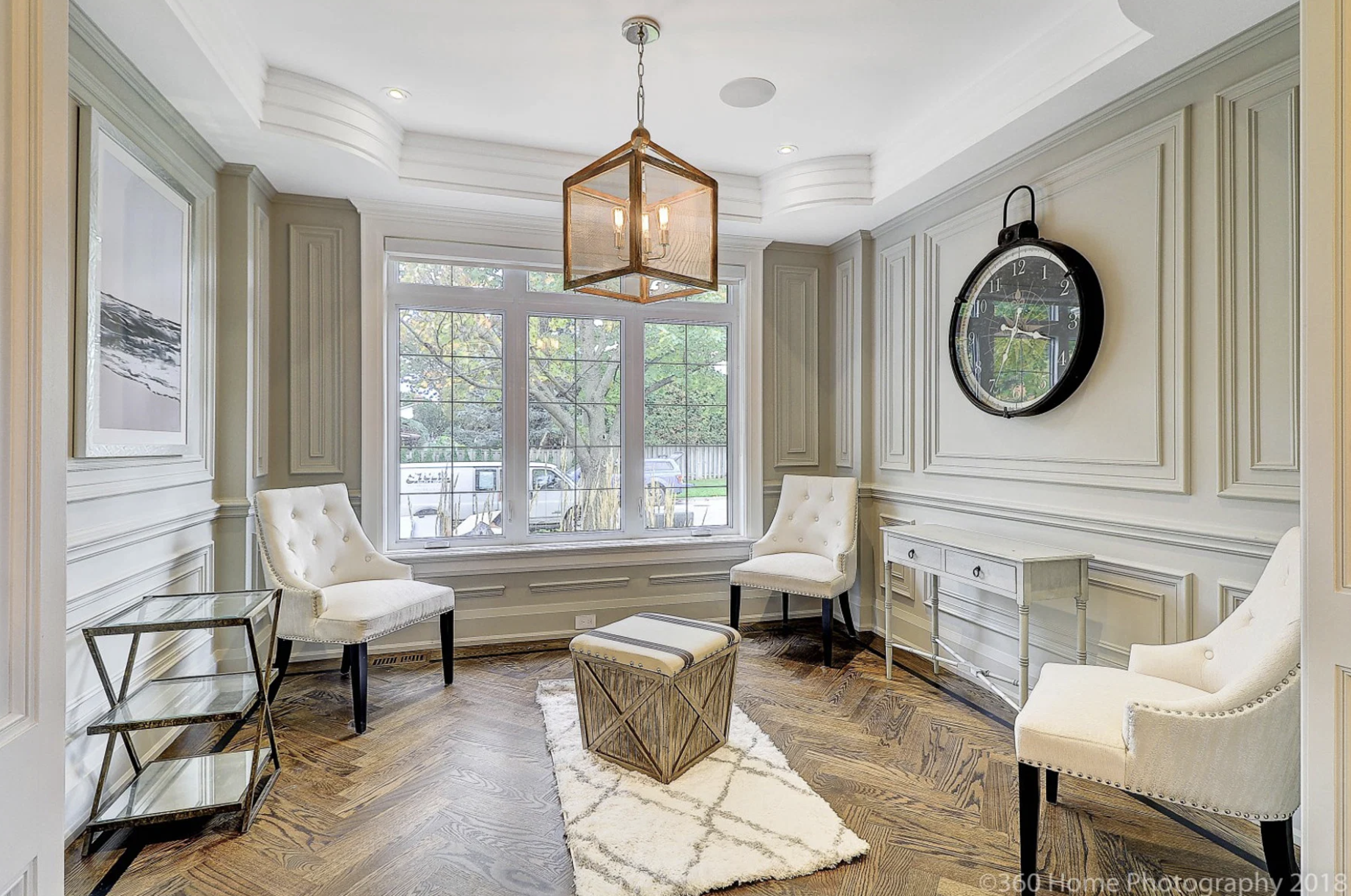 Living room with beige walls, white molding, a large window, two white armchairs, a small white side table, a glass and metal side table, a geometric coffee table on a white rug, a large round wall clock, and a square pendant light.