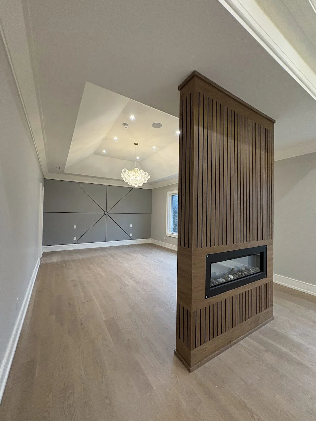 Empty living room with a wood-paneled divider featuring a built-in fireplace, hardwood floors, a modern chandelier, and a large window, with gray wall panels and ceiling lighting.