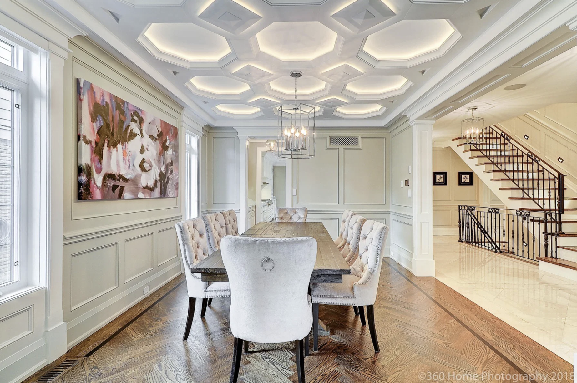 A dining room with a large wooden table and six upholstered chairs. The ceiling has octagonal recessed lighting, and a chandelier hangs above the table. There are large windows flanking a wall with wainscoting, and a staircase with a dark wrought iro
