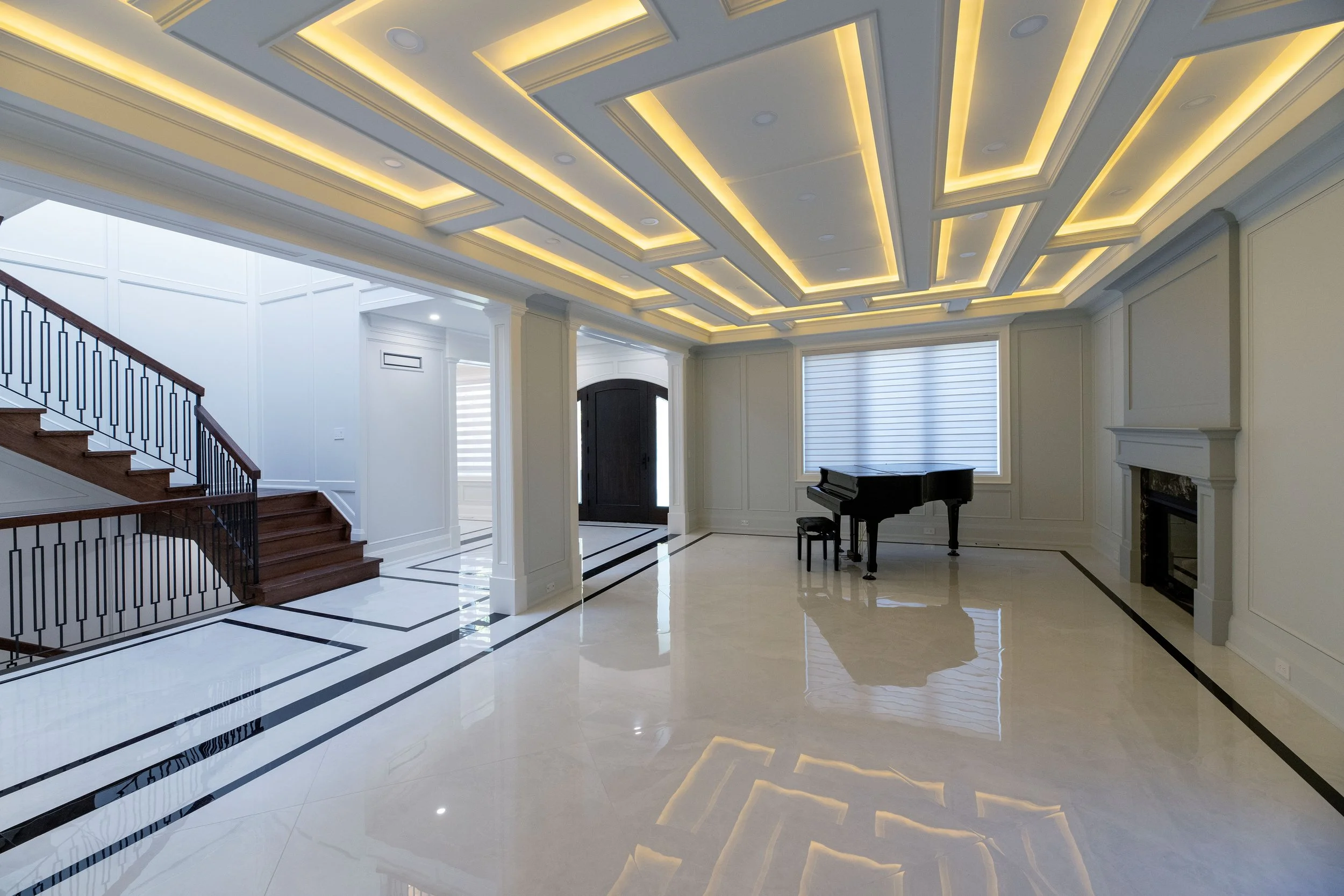 Interior of a luxurious home with a high ceiling featuring recessed lighting, white walls with decorative molding, a staircase with wooden steps and black metal railing, and a black grand piano near large windows.