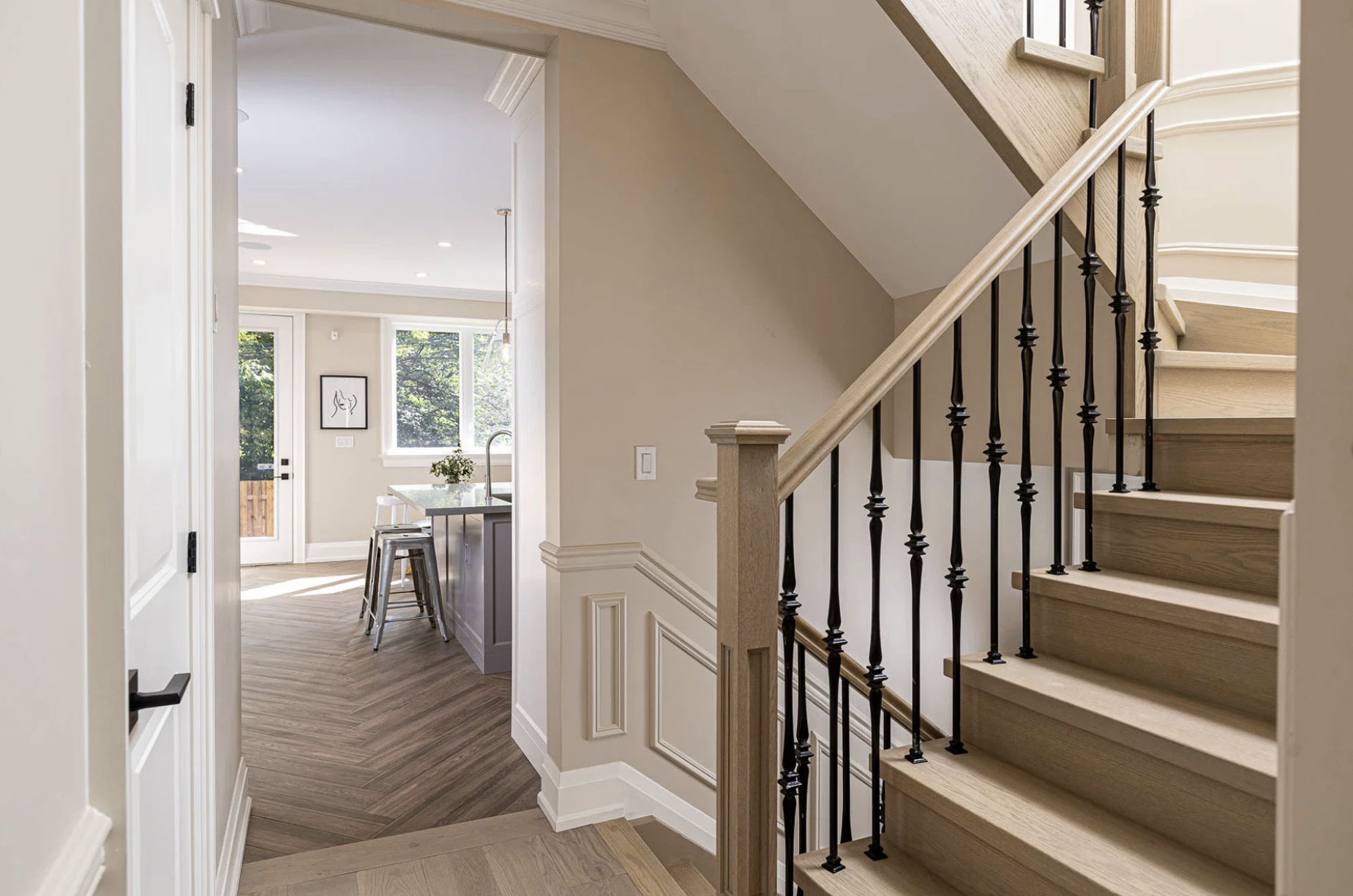 Interior view of a modern home’s stairway with light wooden steps, black balusters, and a beige wall, leading to an open breakfast area with large windows and a glass door with a wooden fence outside.