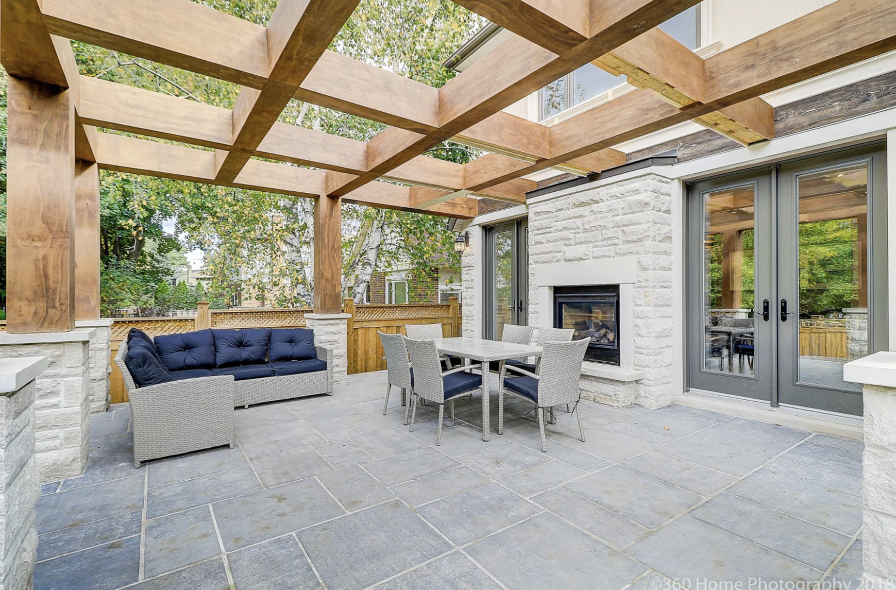Outdoor patio with grey tiled flooring, a cushioned outdoor sofa, a dining table with six chairs, a fireplace, wooden pergola framework, and sliding glass doors leading into the house, with trees in the background.