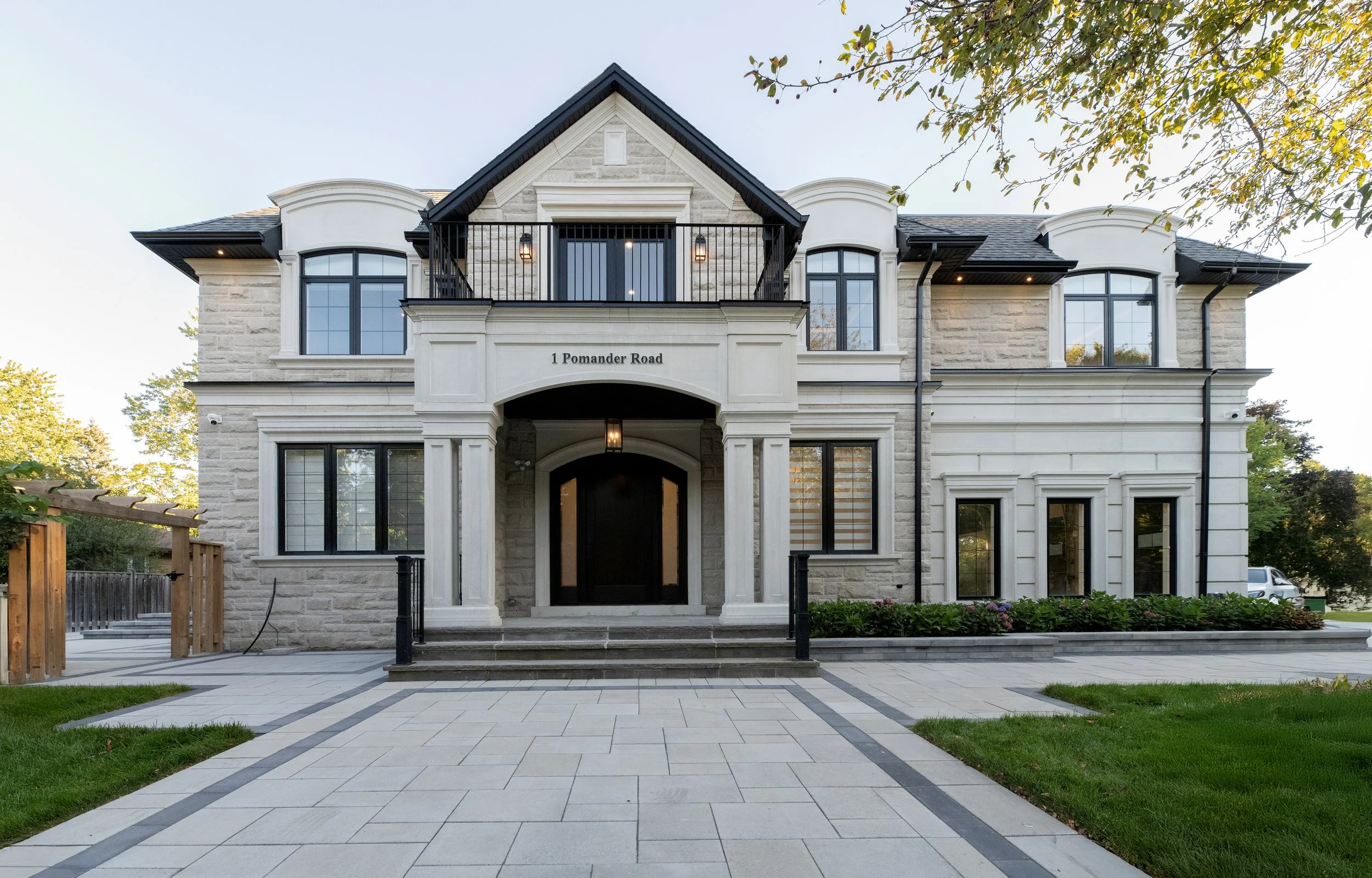 Front view of a modern two-story house with an arched entryway, white stone facade, and large black-framed windows, surrounded by a landscaped yard with a paved walkway.