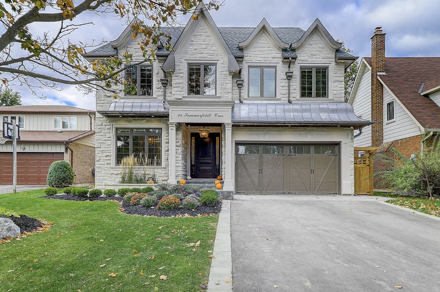 A modern white two-story house with a landscaped front yard, grey garage door, and decorative pumpkins on the steps, featuring a sign with the house address '34 Summerfield Cres.'