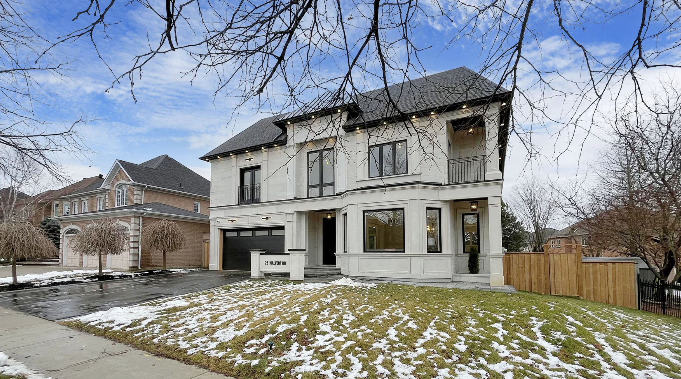 A modern three-story house with a black garage door, large windows, and a front porch, located at 251 Calvert Road. The front yard has a sidewalk, patches of snow, and leafless trees, with neighboring houses visible in the background under a partly c