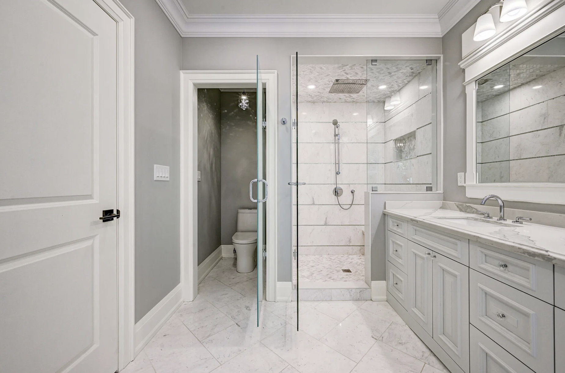 Bathroom with gray walls, white doors, marble vanity, mirrored cabinet, glass-enclosed shower with marble tiles, and a separate toilet area.